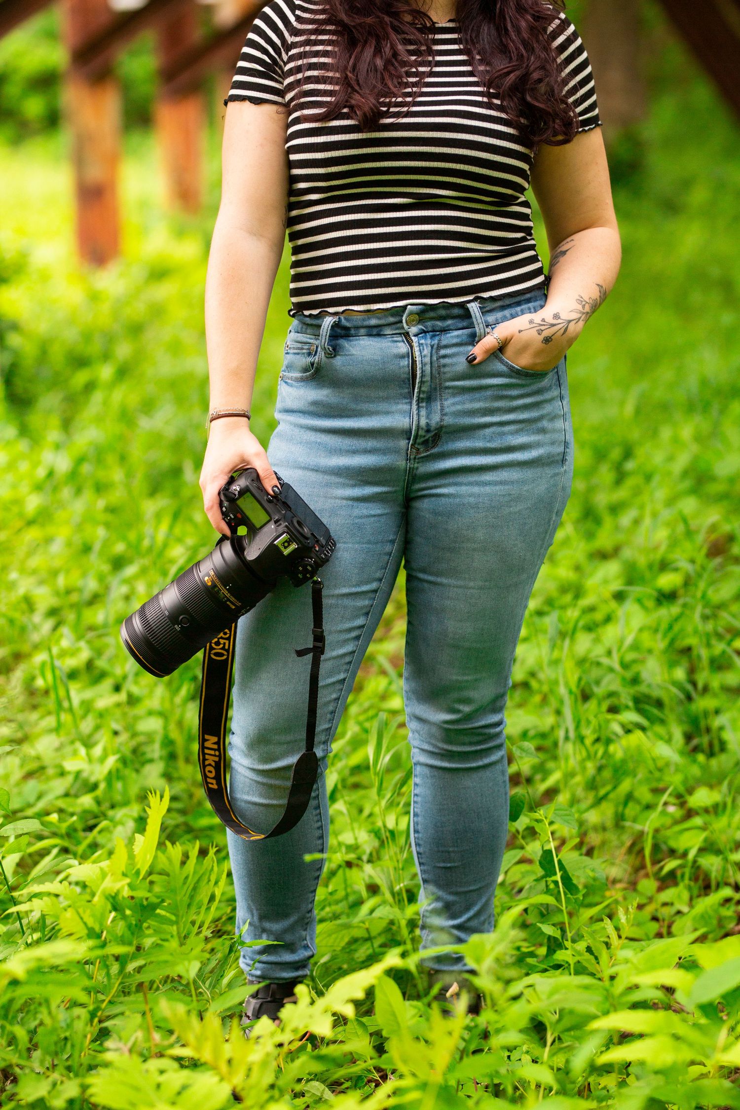 Person in striped shirt and jeans holding camera while standing in green field.
