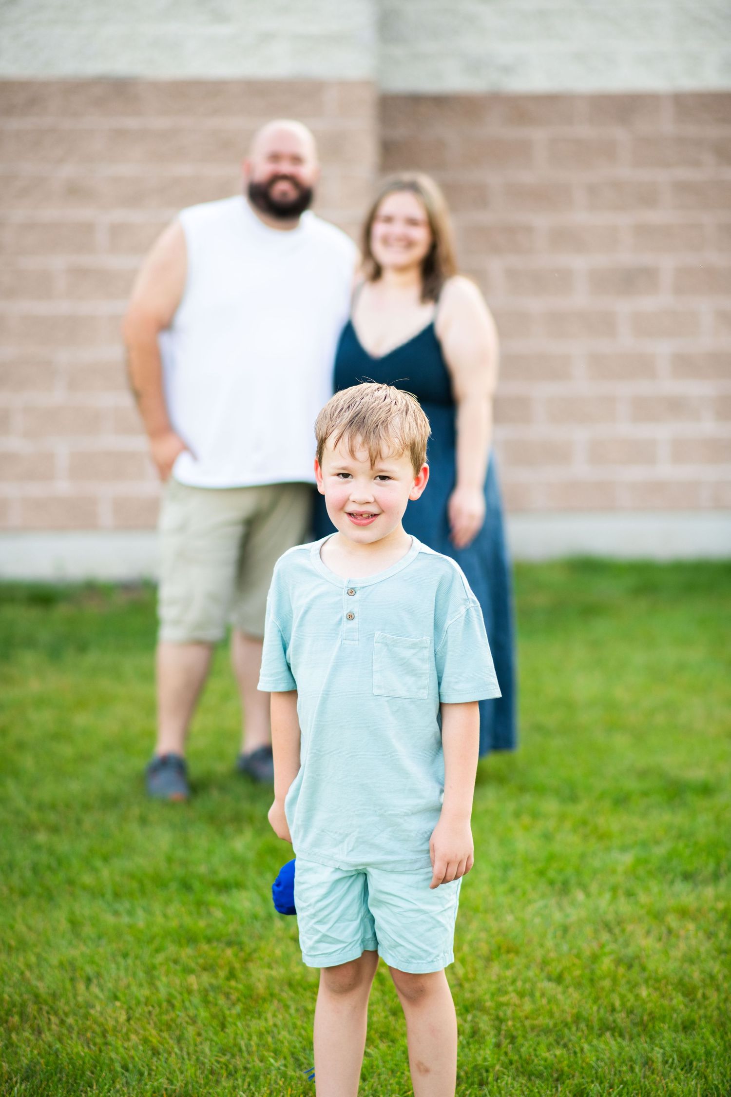 Child in light blue outfit playing and smiling on grass with brick wall background.