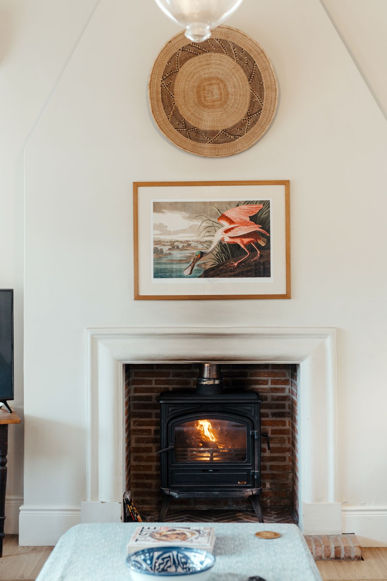 A cozy fireplace with a lit wood stove, framed Audubon bird print, and circular woven wall decor in a modern living room.