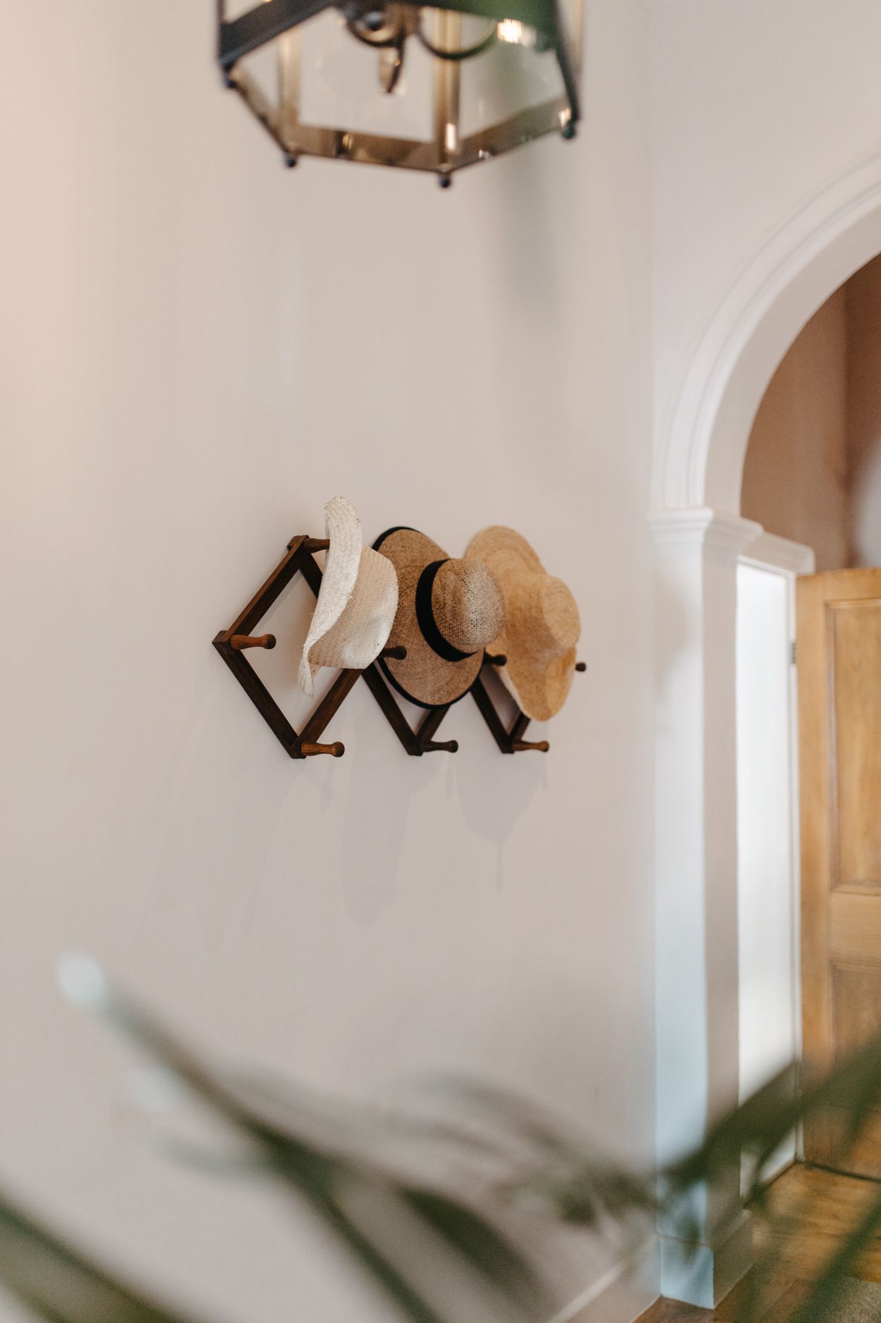 Straw hats hanging on a wooden accordion wall rack beneath a vintage pendant light in a bright hallway.