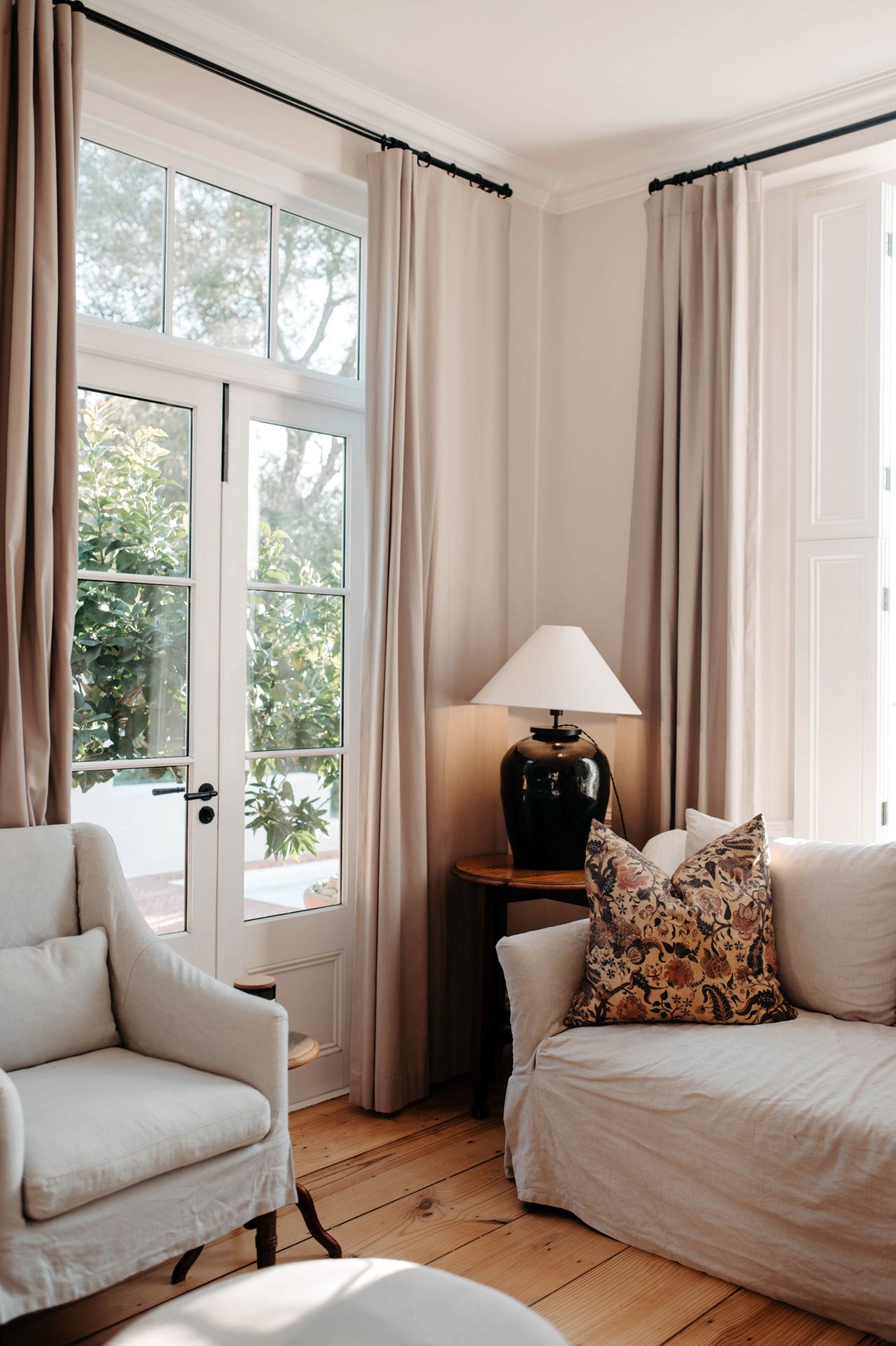Bright and airy living room with white slipcovered armchairs, hardwood floors, and large windows with beige curtains.