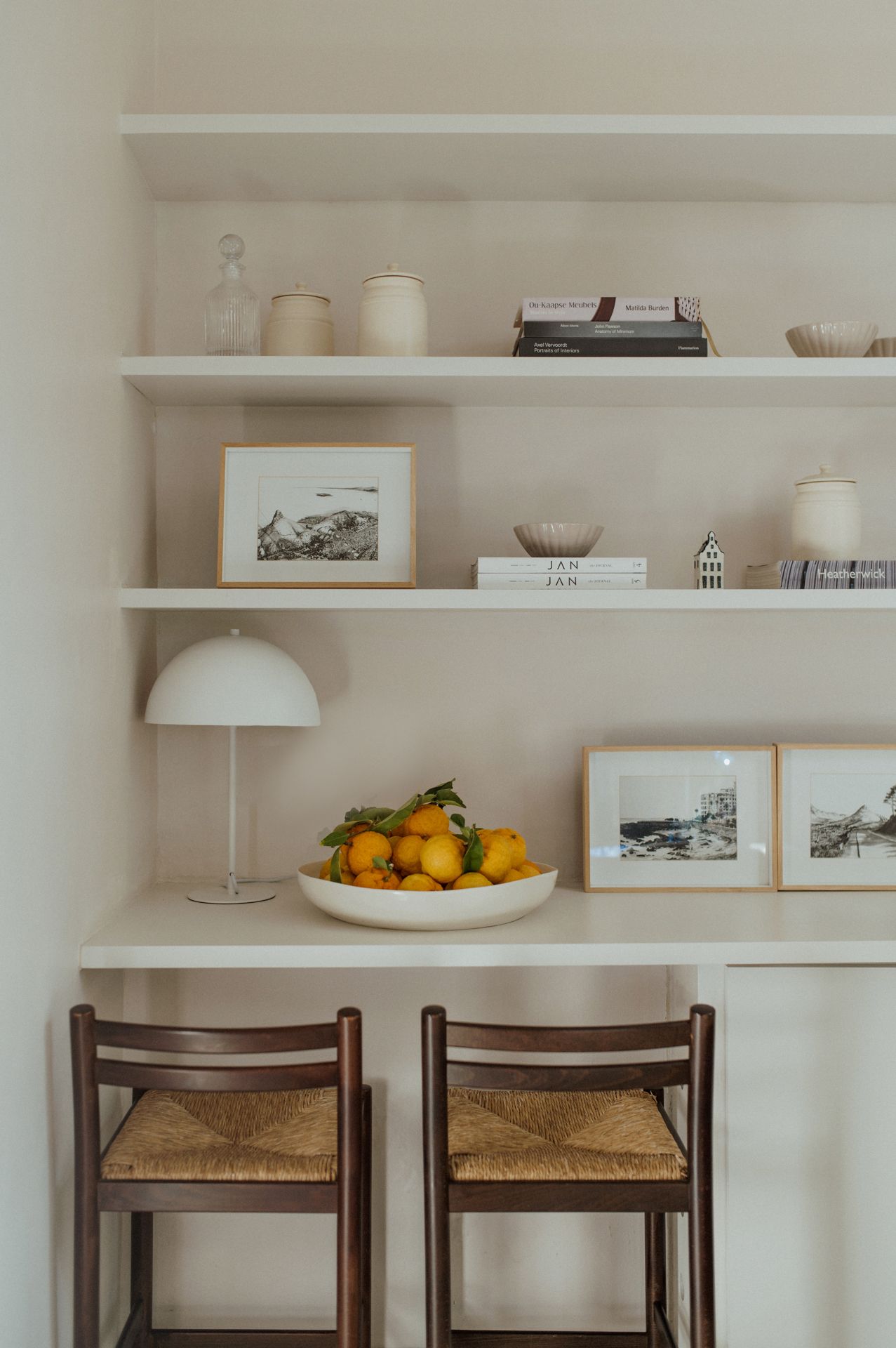 White floating shelves display artwork and decor above wooden chairs with woven seats in a minimalist interior space.