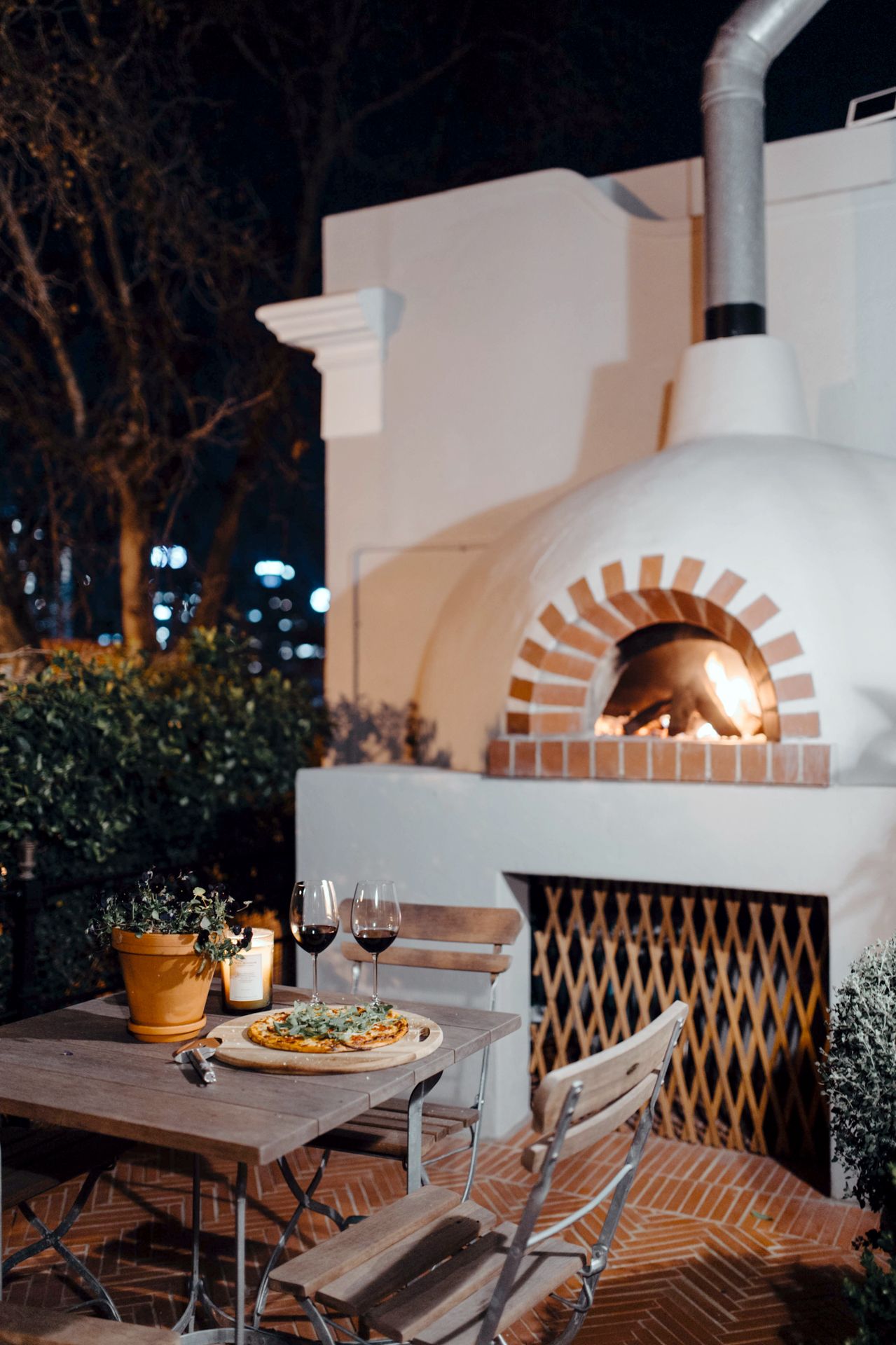 A cozy outdoor patio with a brick pizza oven and wine glasses on wooden tables illuminated in warm evening light.