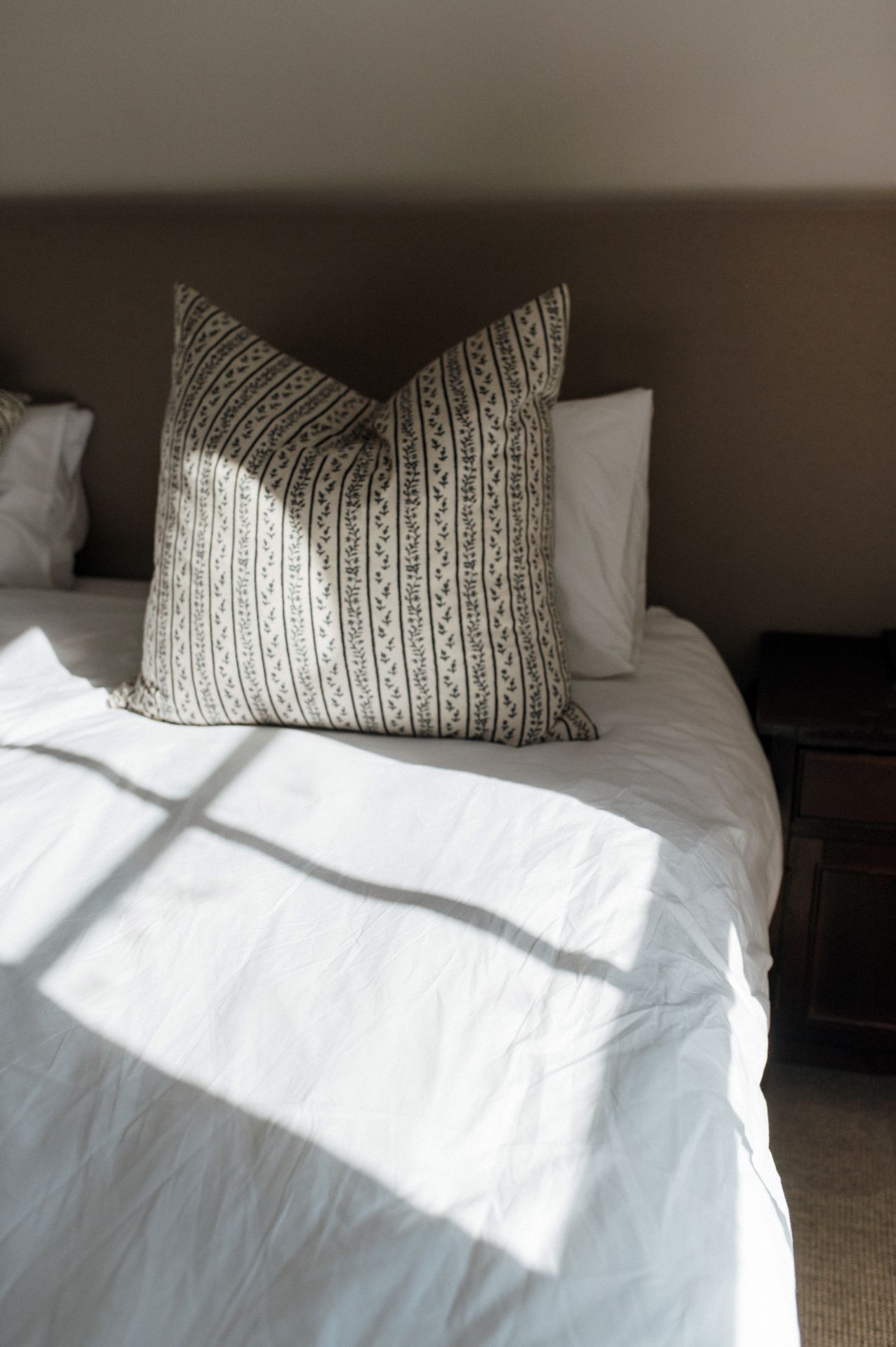 Sunlight casts dramatic shadows across a white bed with a decorative patterned pillow in neutral tones.