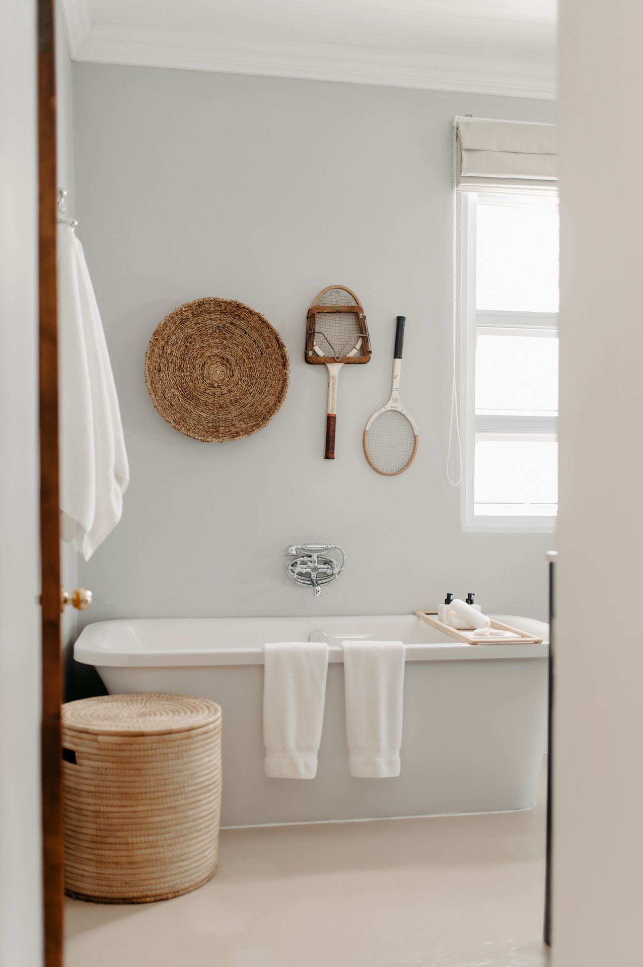 Minimalist white bathroom with vintage decor featuring a freestanding tub, woven basket, and natural accents on light gray walls.