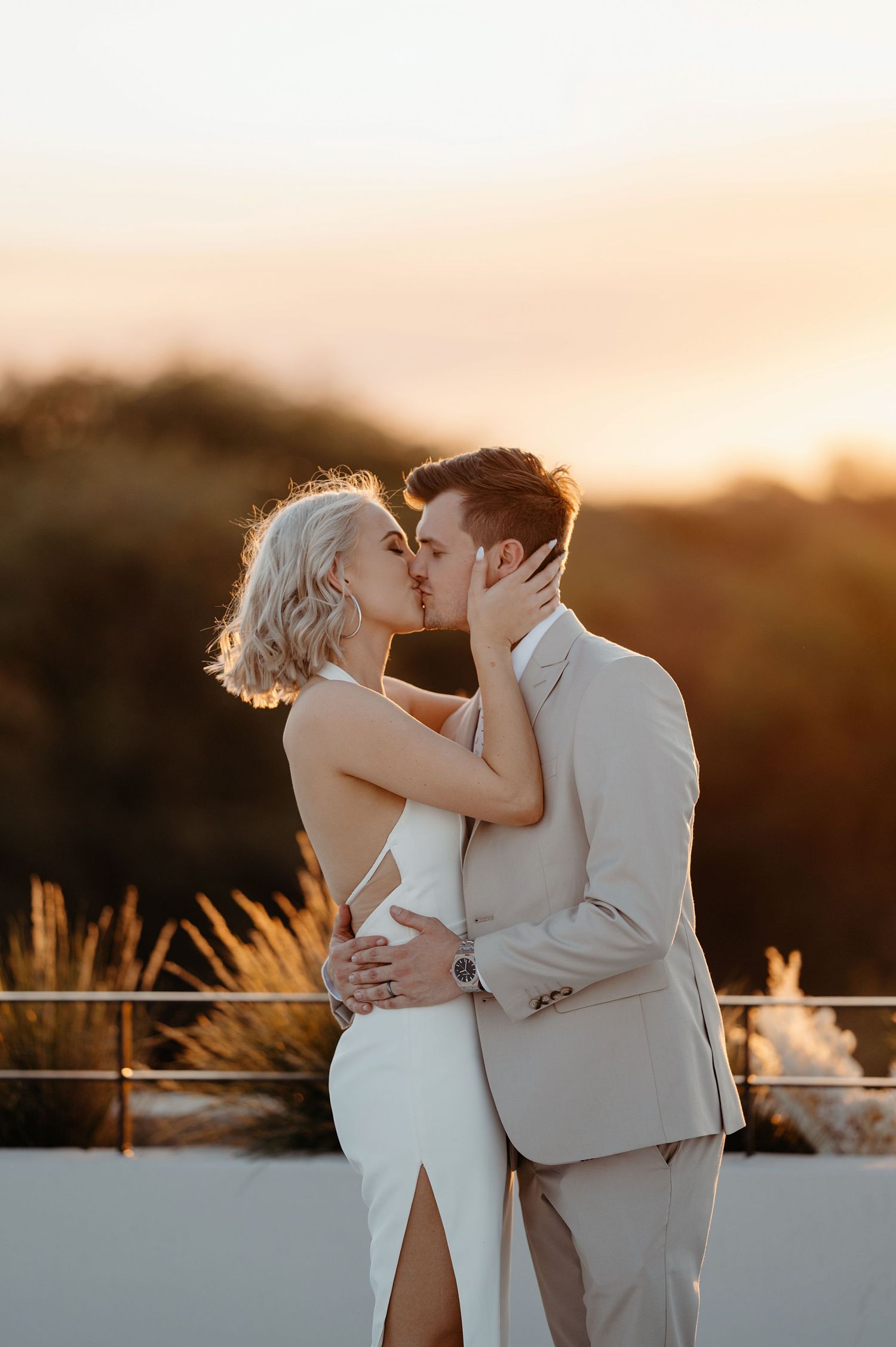 A couple shares a romantic kiss at sunset on a rooftop terrace with warm golden lighting.