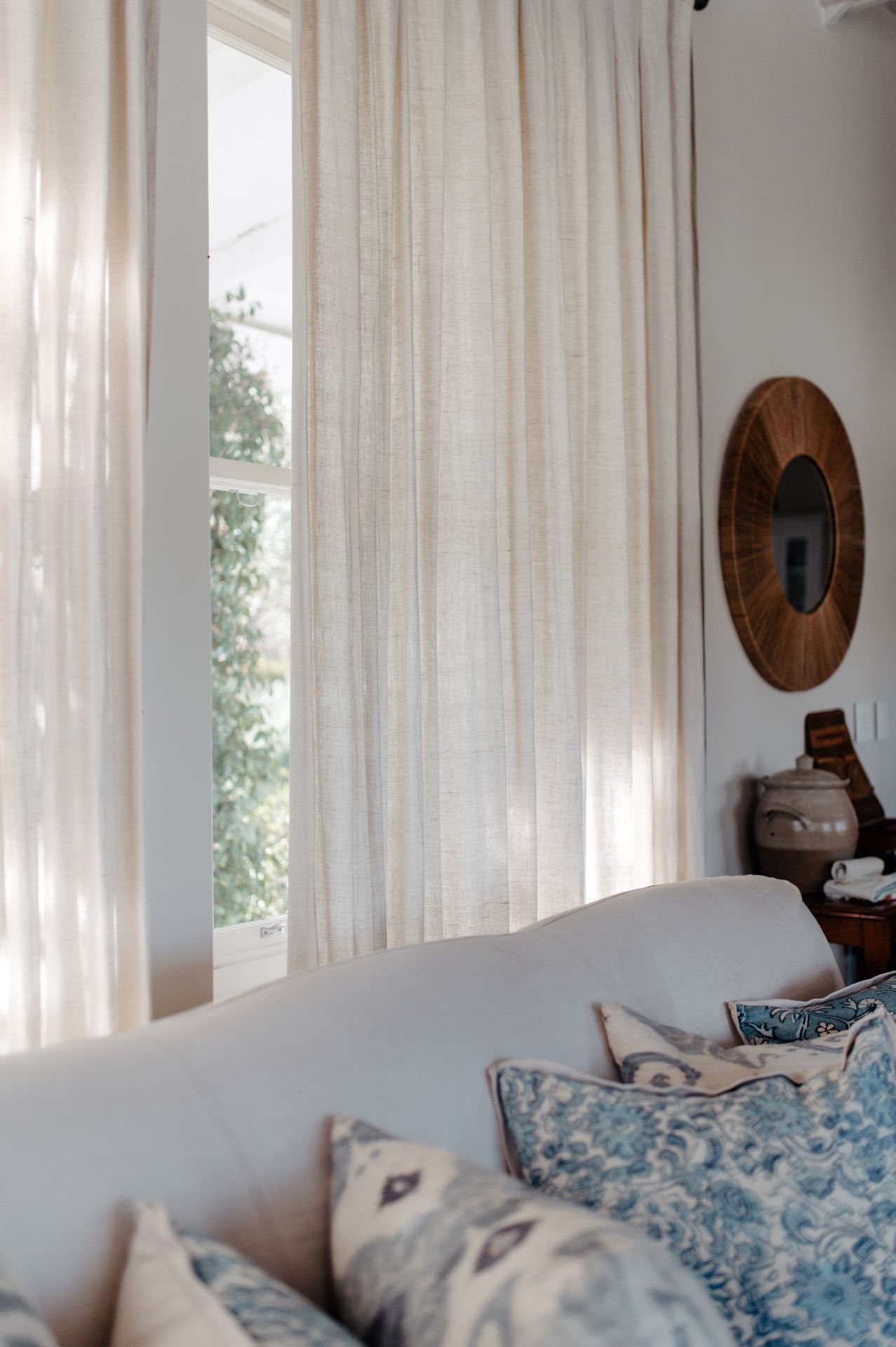 Sunlit living room with sheer white curtains, blue patterned throw pillows on a light gray couch, and wooden wall decor.