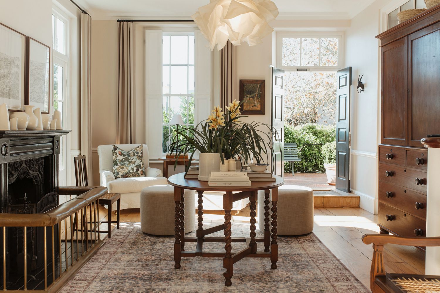 Bright living room with vintage furniture, beige ottomans, wooden table, and large windows bringing in natural light.