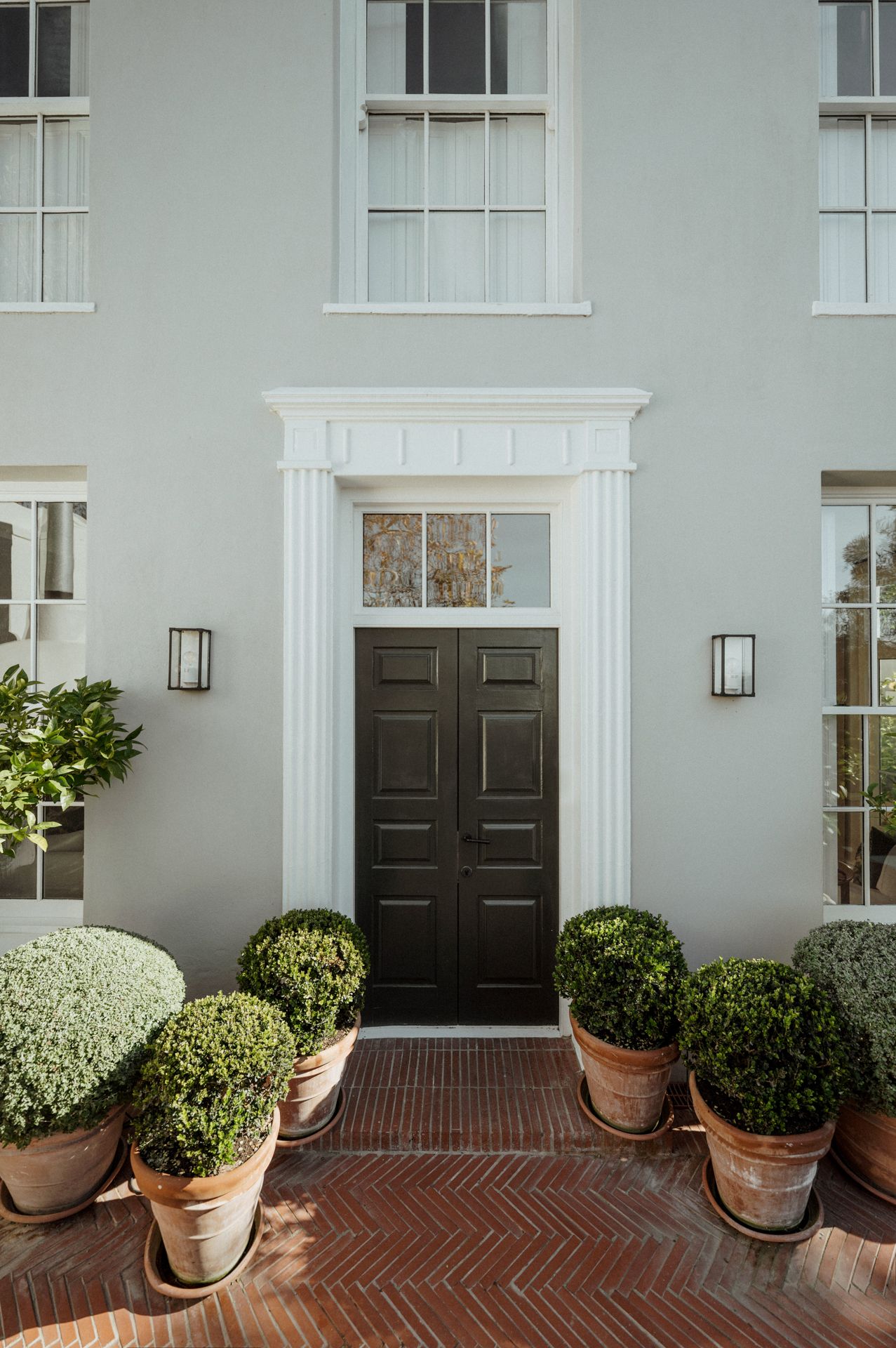 Elegant black front doors with white trim on a gray building facade, flanked by potted topiary plants on herringbone brick.