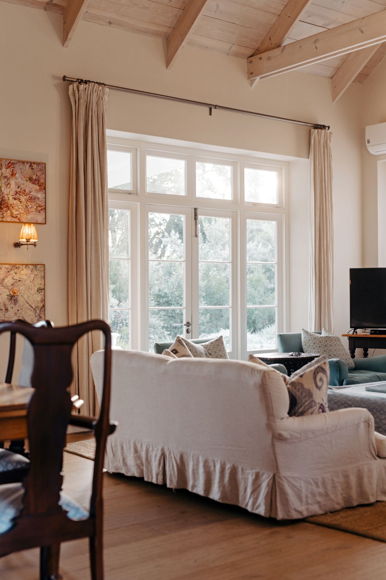 Modern living room with vaulted wood beam ceiling, white slip-covered furniture, and large windows letting in natural light.