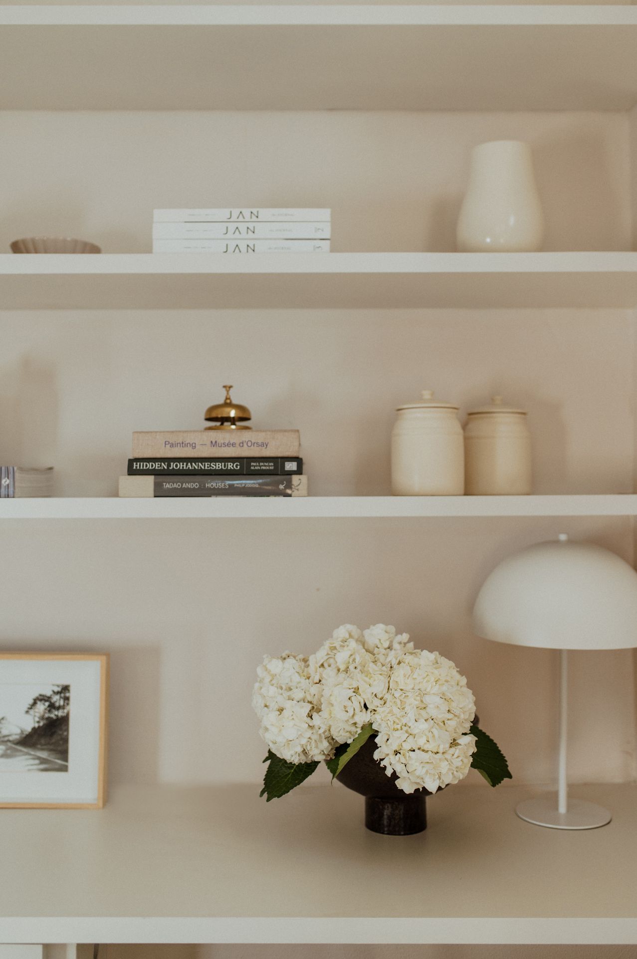 Minimalist white floating shelves display books, decorative objects, and hydrangea flowers in a neutral toned room.