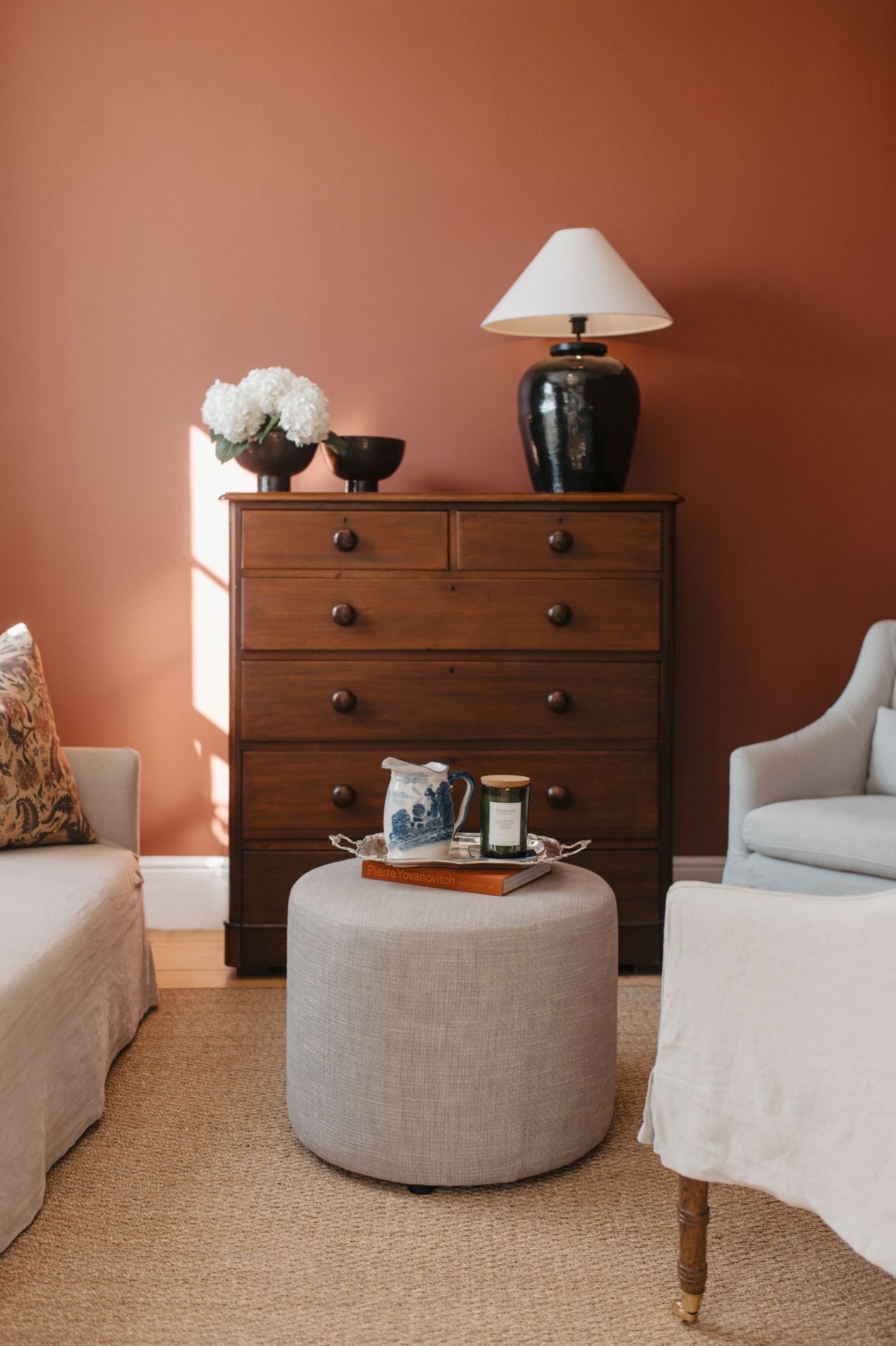 A minimalist living room with white upholstered seating, wooden dressers, and decorative objects against a terracotta wall.