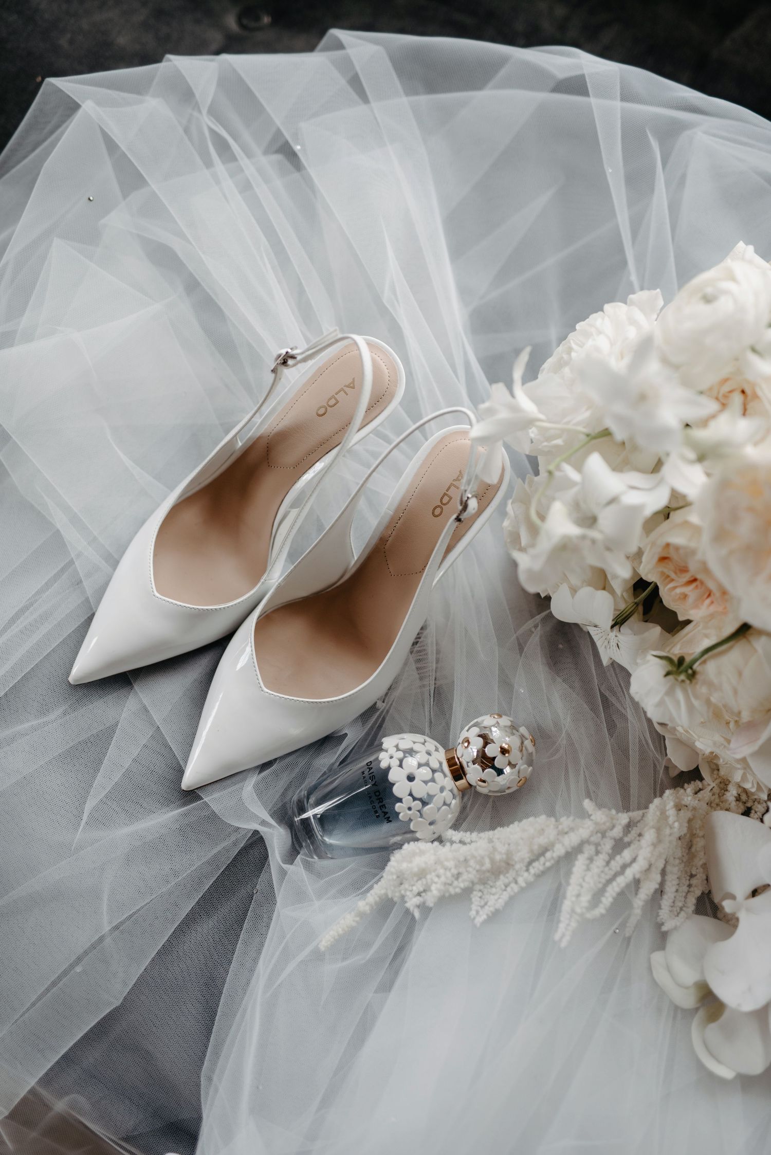 Elegant white bridal heels and delicate tulle veil arranged with soft white floral bouquet in romantic wedding styling.