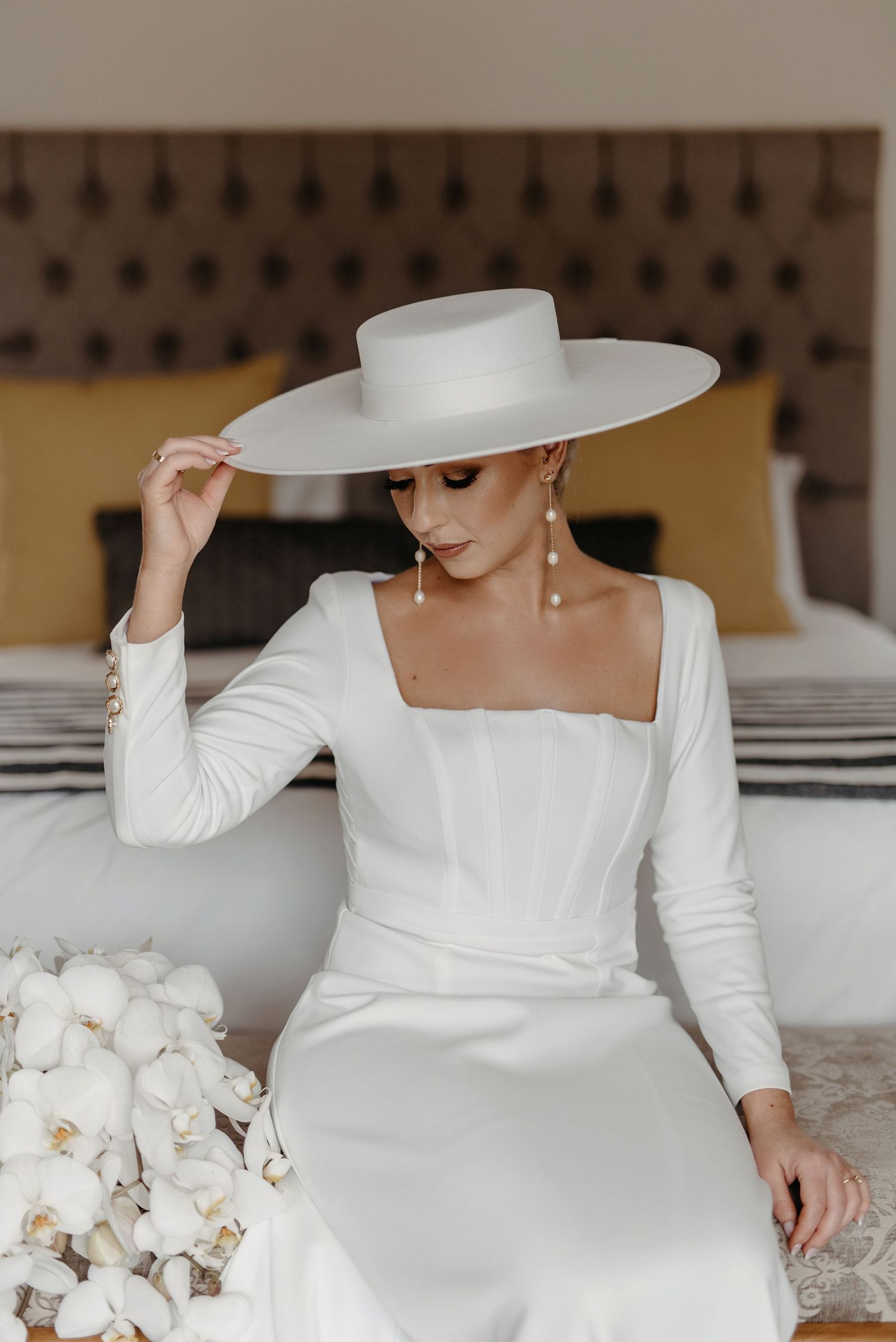 Elegant bridal fashion featuring a white long-sleeved dress and wide-brimmed hat with delicate orchids in the scene.