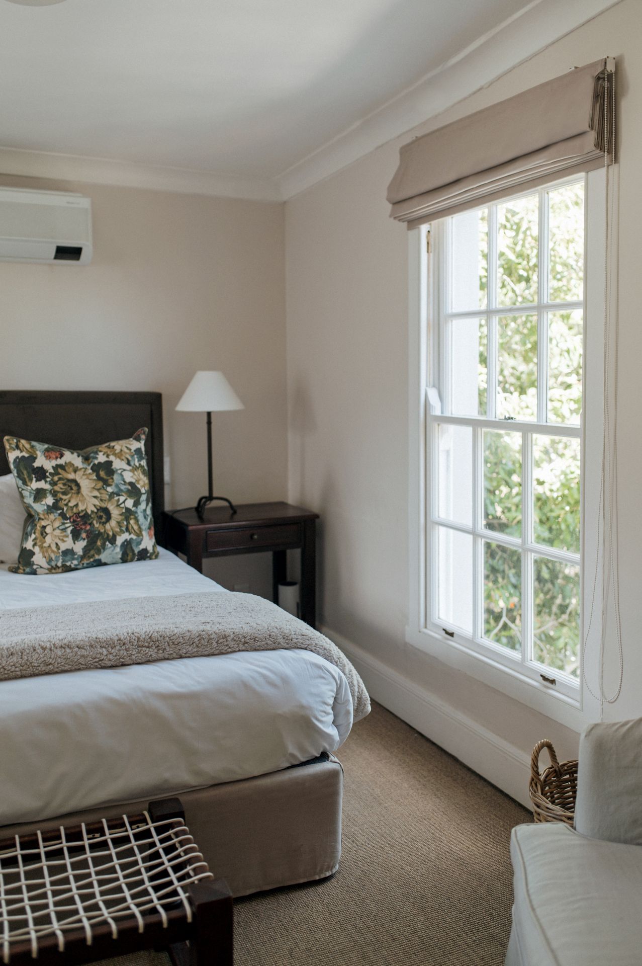 A cozy bedroom with a large window, floral throw pillow, and beige decor featuring natural light and carpeted floors.
