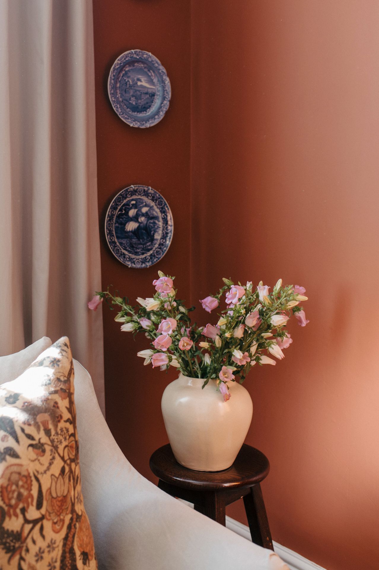 White ceramic vases with pink and white flowers sit atop tables against a terracotta wall with decorative blue plates.