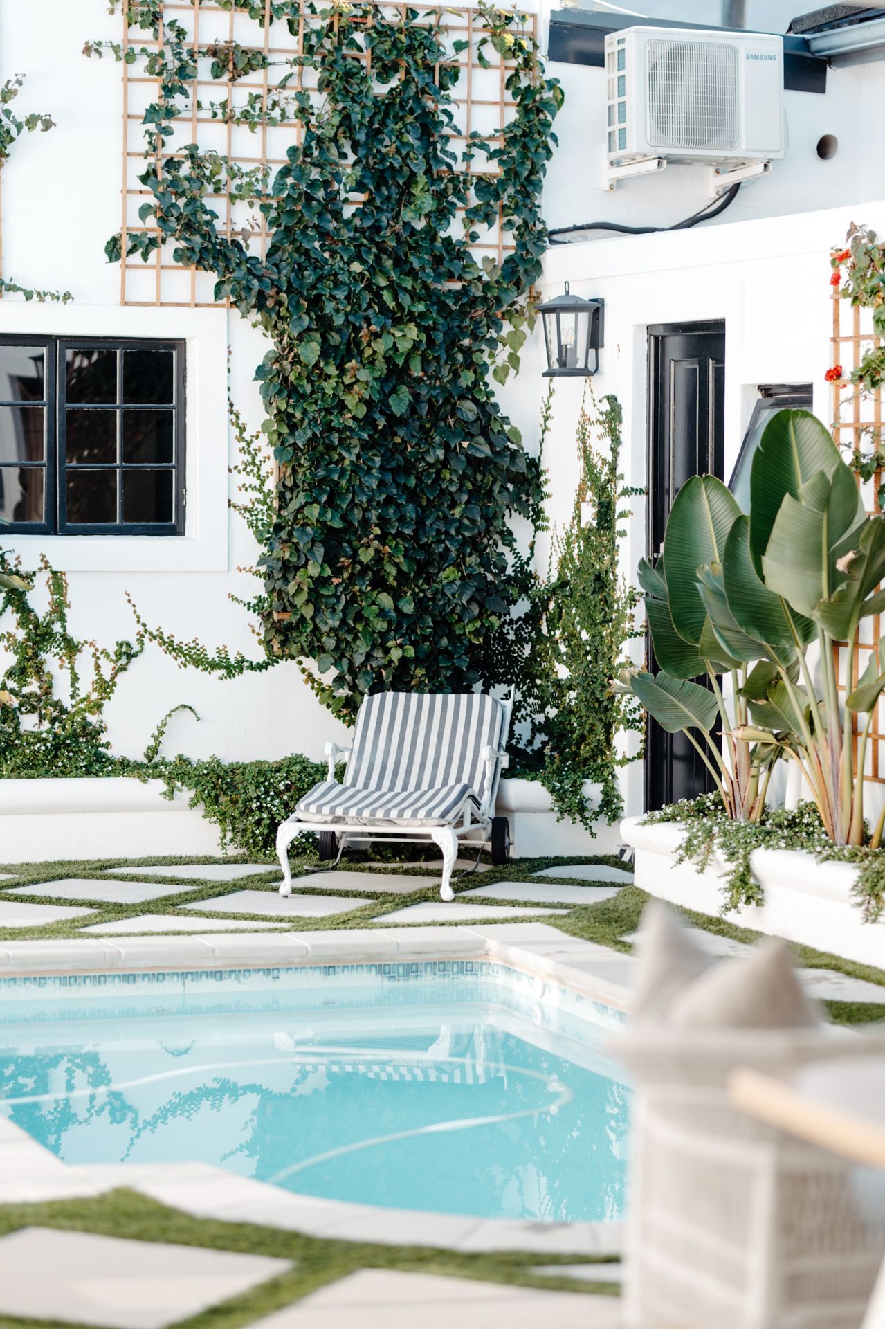Modern backyard pool area with white walls, climbing vines, and striped lounge chairs creates a serene outdoor oasis.