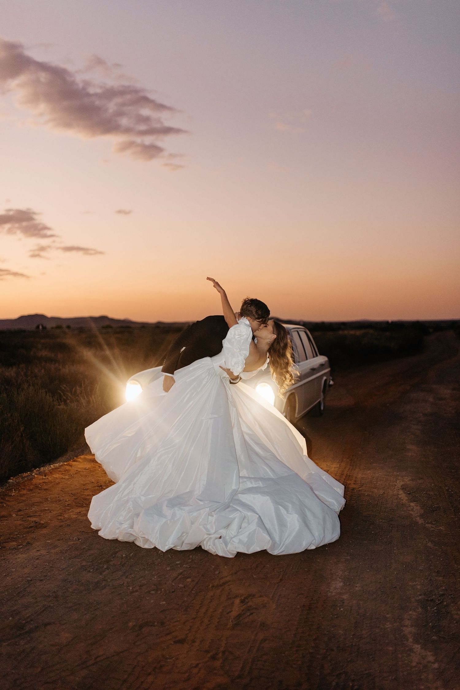 Dramatic sunset silhouette of couple in wedding attire near car headlights on dirt road.