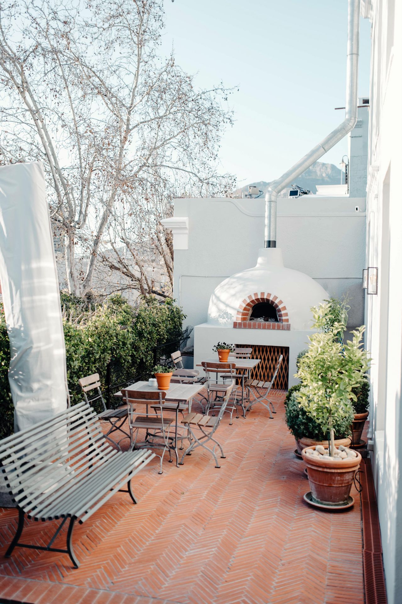 A Mediterranean-style terrace with herringbone brick flooring, white walls, and wooden benches features a built-in brick oven.