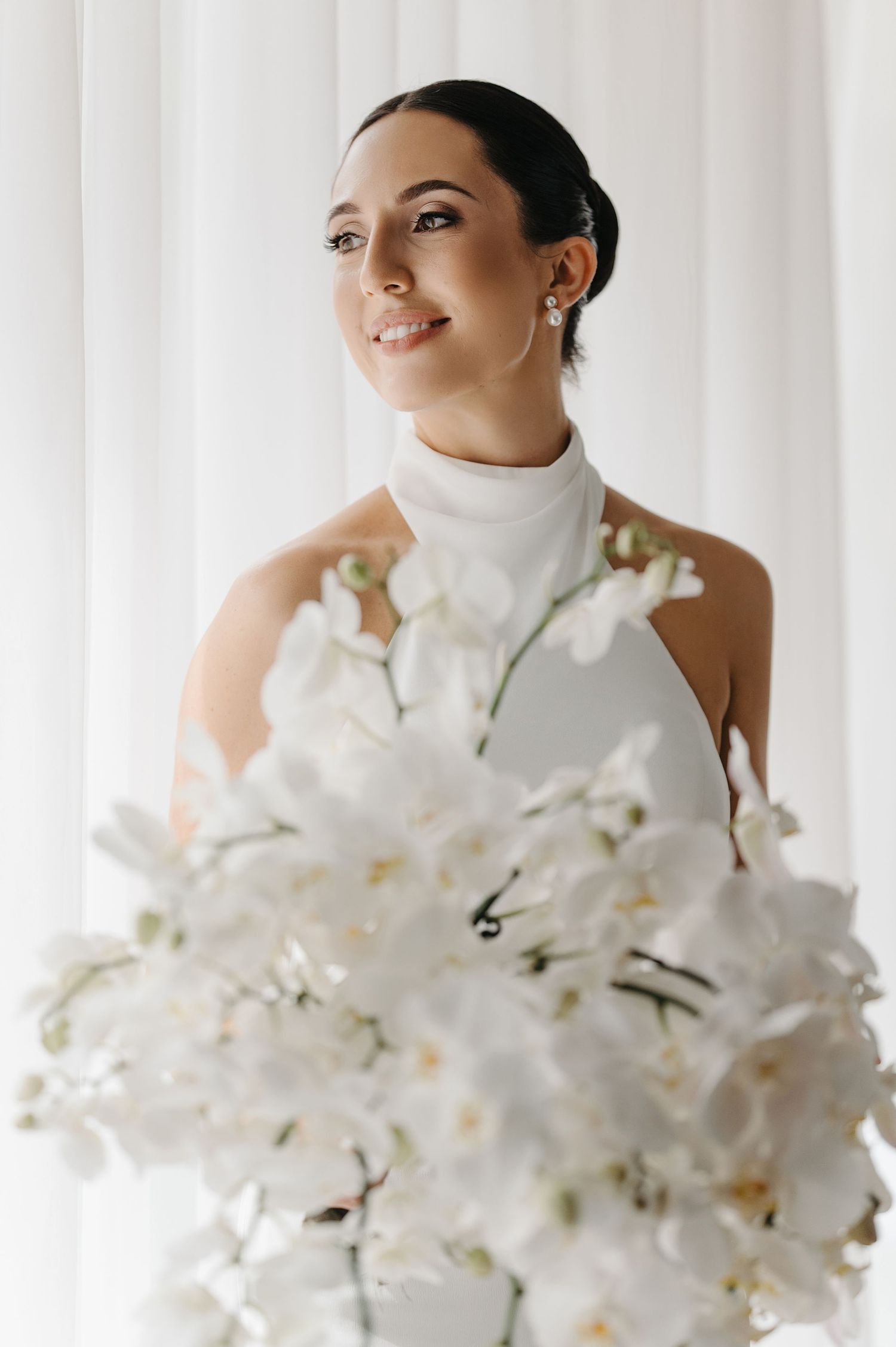 Person in white turtleneck poses with elegant white orchid bouquet against light curtain background.