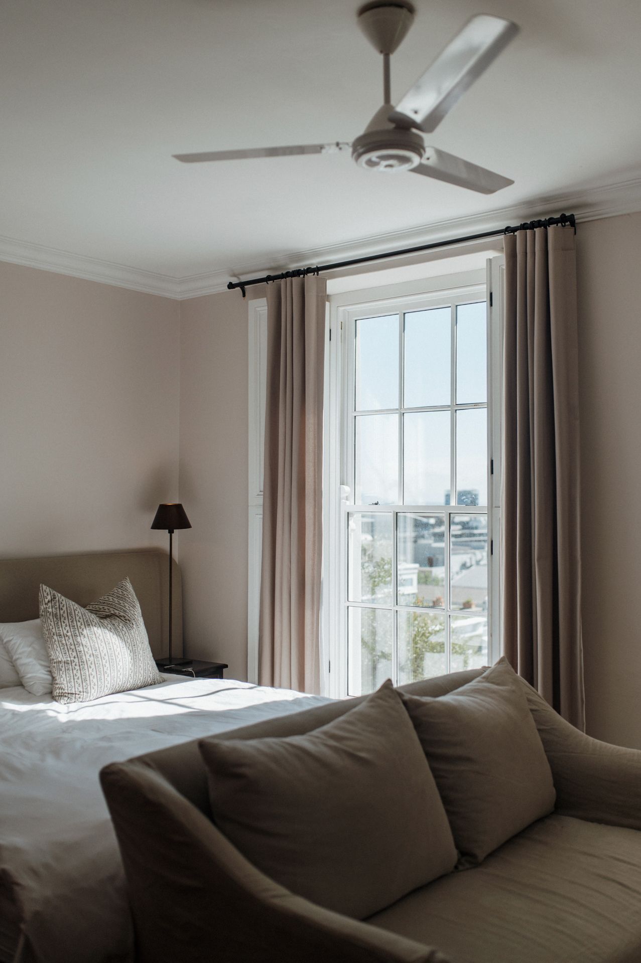 Modern bedroom interior with ceiling fan, large windows, gray curtains, and comfortable bedding in neutral tones.