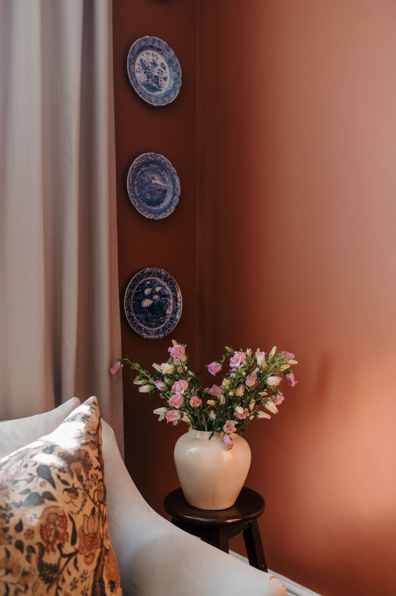 Decorative blue and white plates hang on a terracotta wall above a white vase with pink flowers.