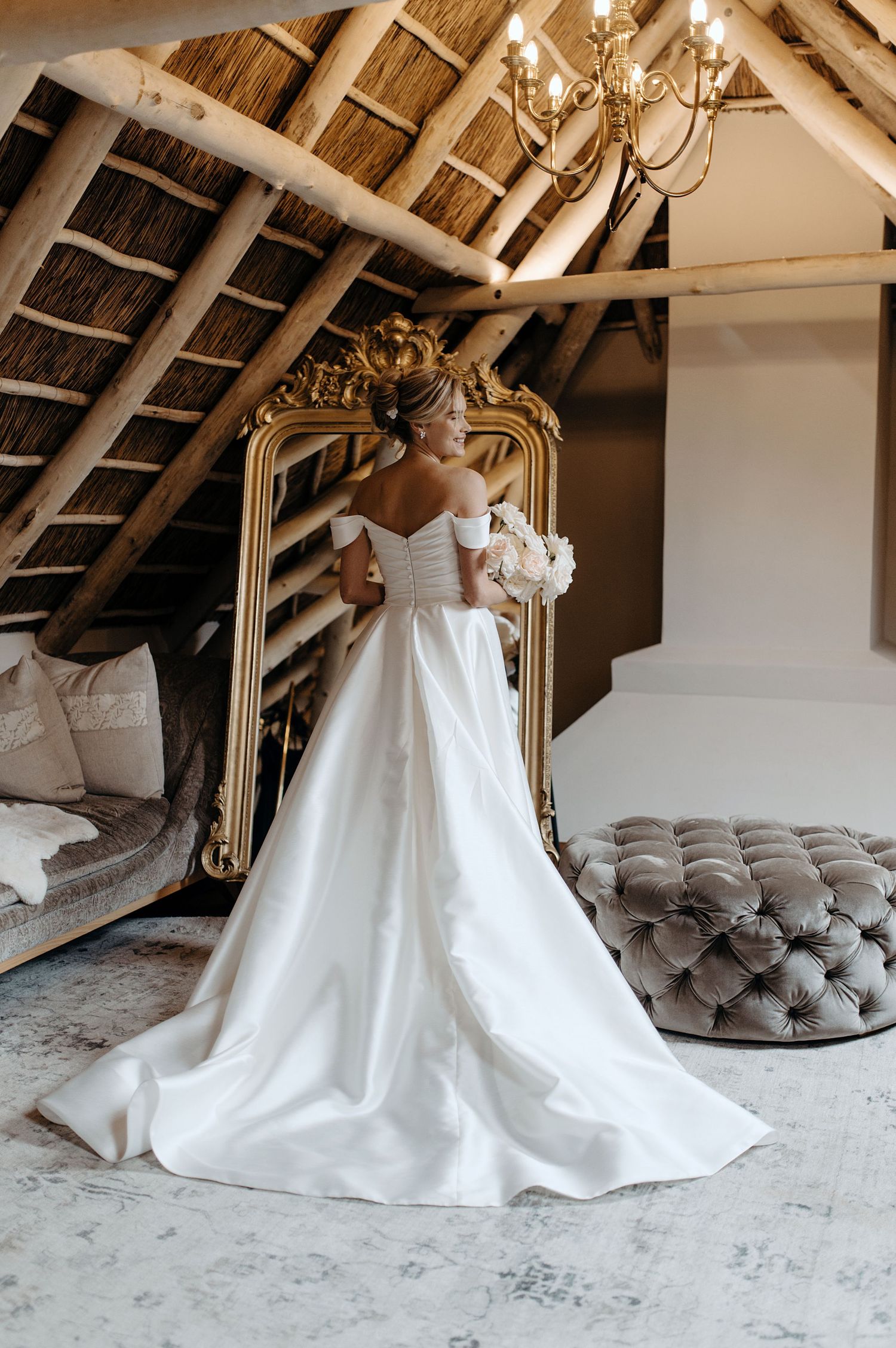 Elegant white wedding dress with a full skirt displayed in a rustic attic setting with exposed wooden beams and chandelier.