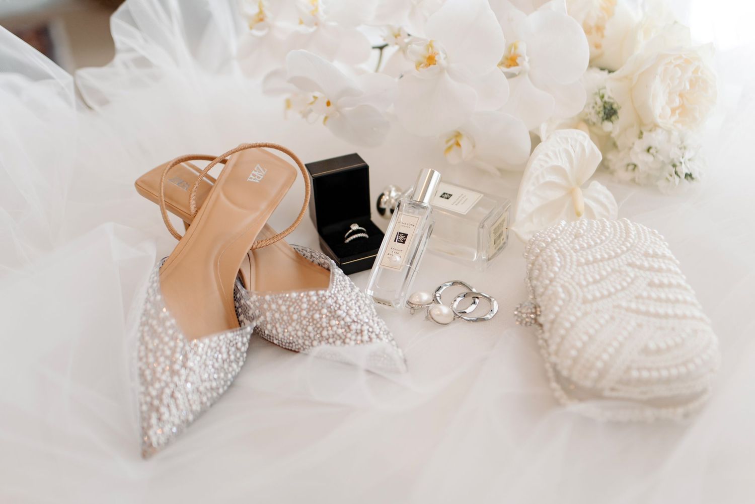 Elegant bridal accessories including sparkly pointed heels, wedding rings, and a white knit beanie arranged on tulle fabric.