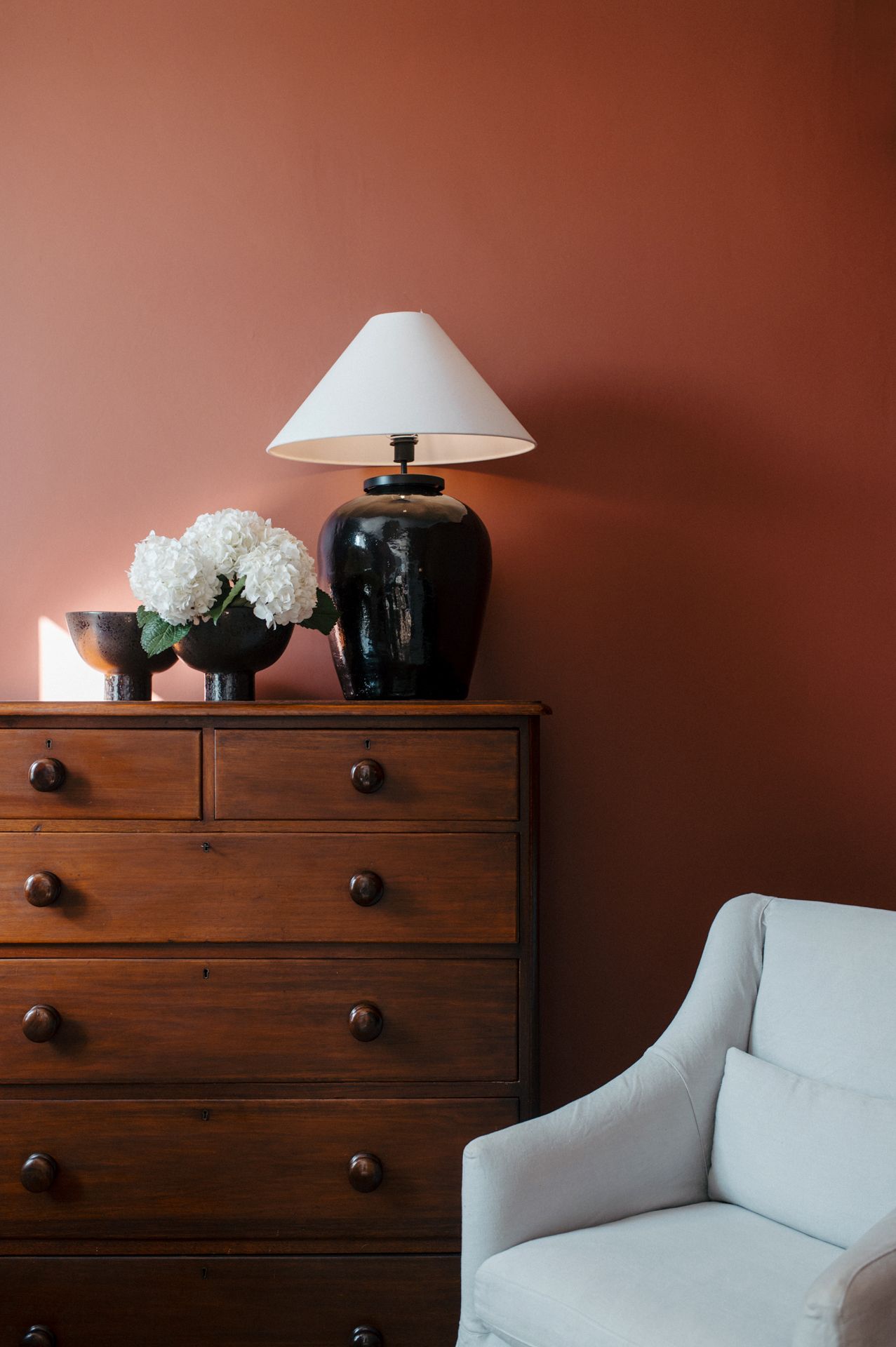 Wooden dresser with black knobs against a brown wall, decorated with a white lamp and decorative objects.