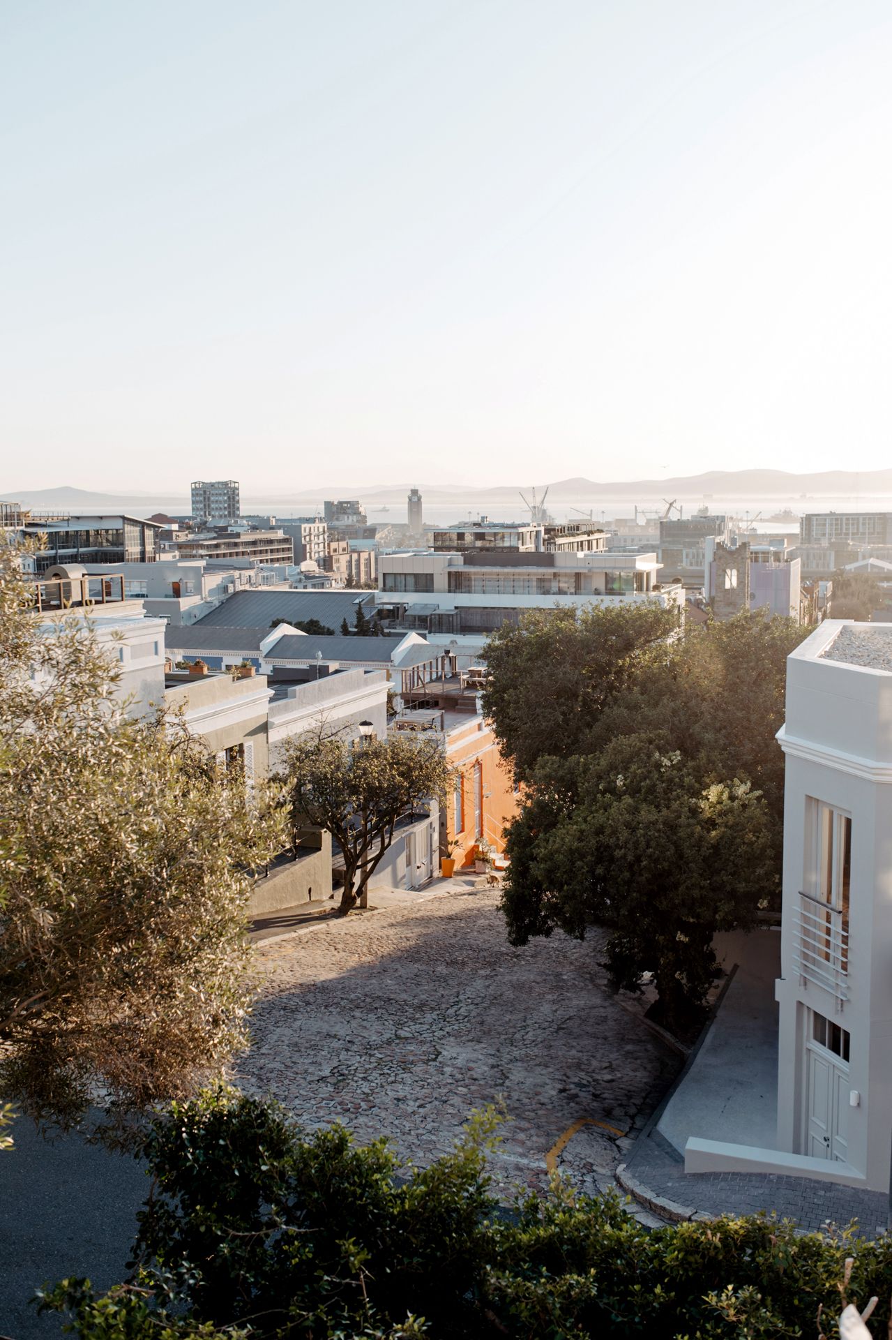 Elevated view of a Mediterranean cityscape with white buildings and trees against a hazy sky during golden hour.