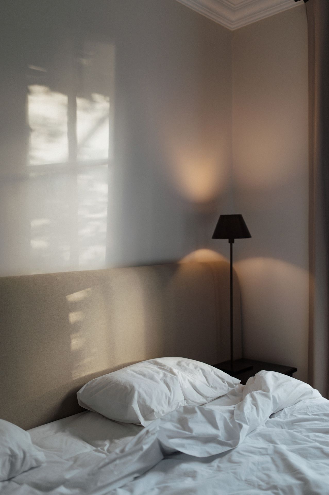 A messy unmade bed with white sheets and a black lamp in a sunlit bedroom corner.