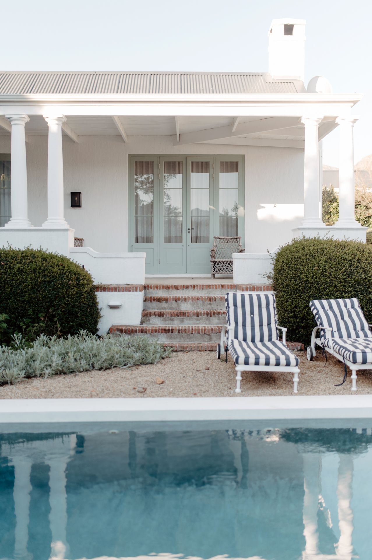 Luxurious poolside setting with striped lounge chairs and manicured hedges against a white Mediterranean-style home.