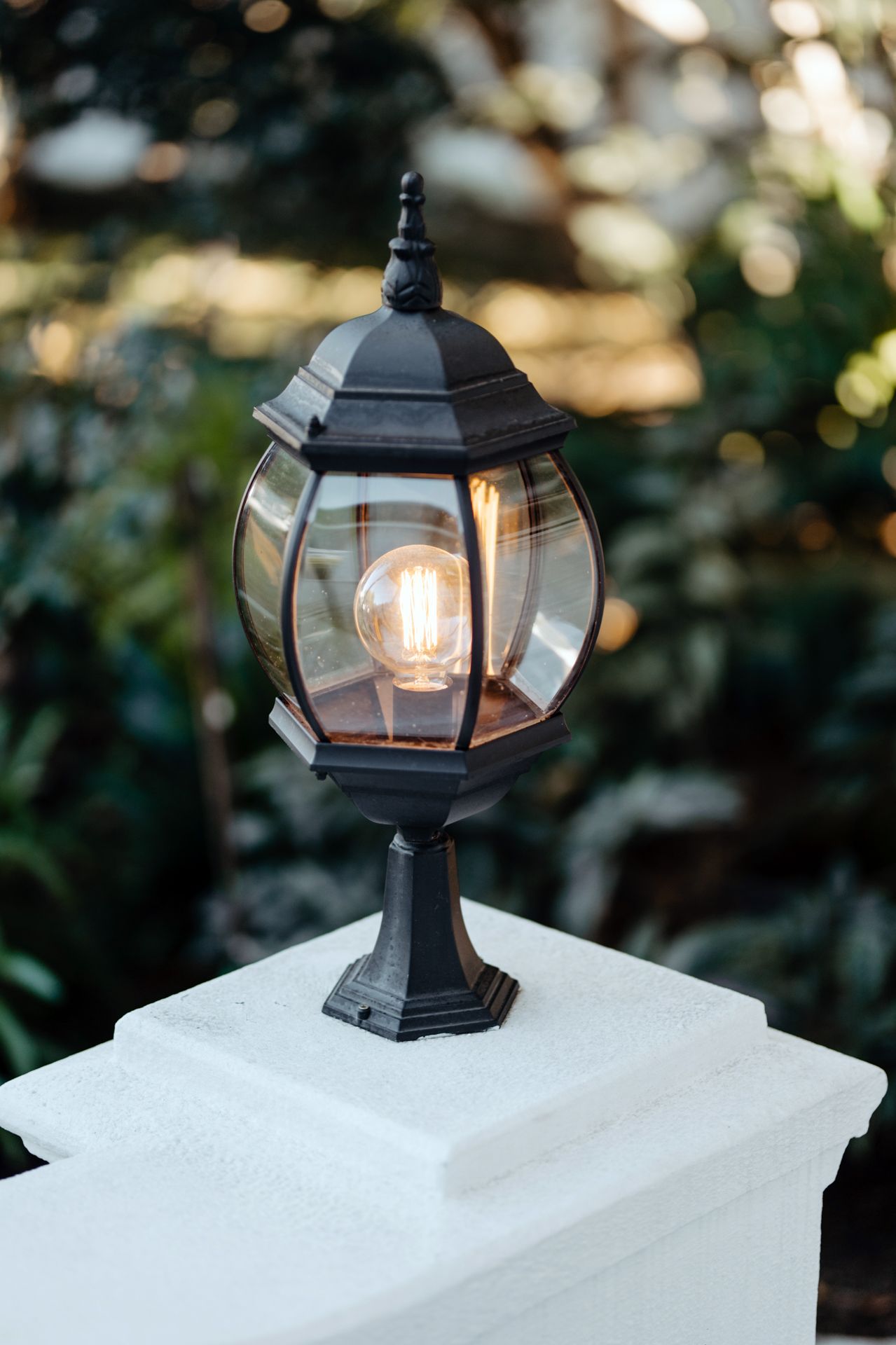 Black outdoor lantern light fixture mounted on white post against blurred garden background.