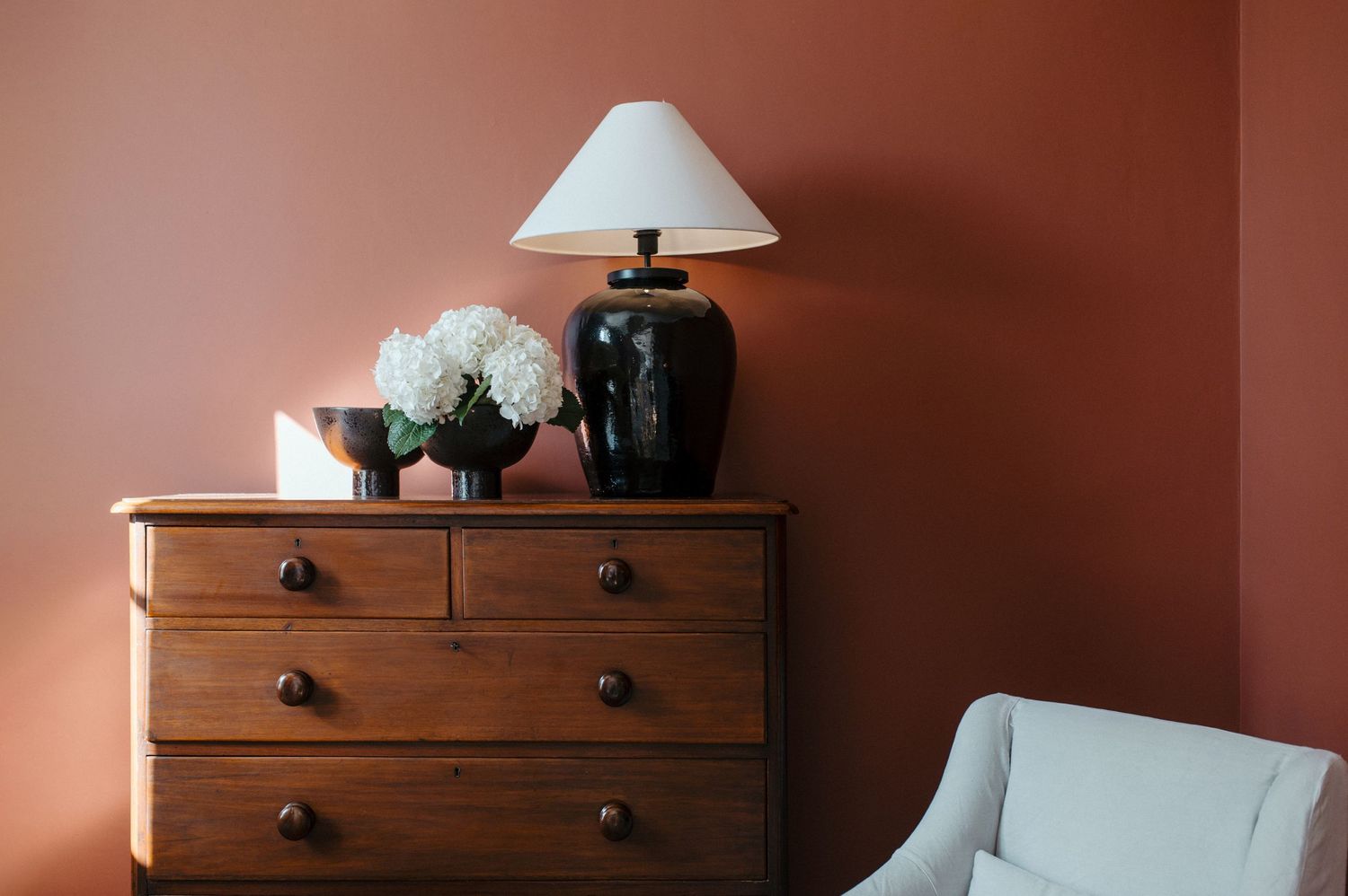 A wooden dresser with black lamp and white flowers against a terracotta wall next to a light-colored armchair.