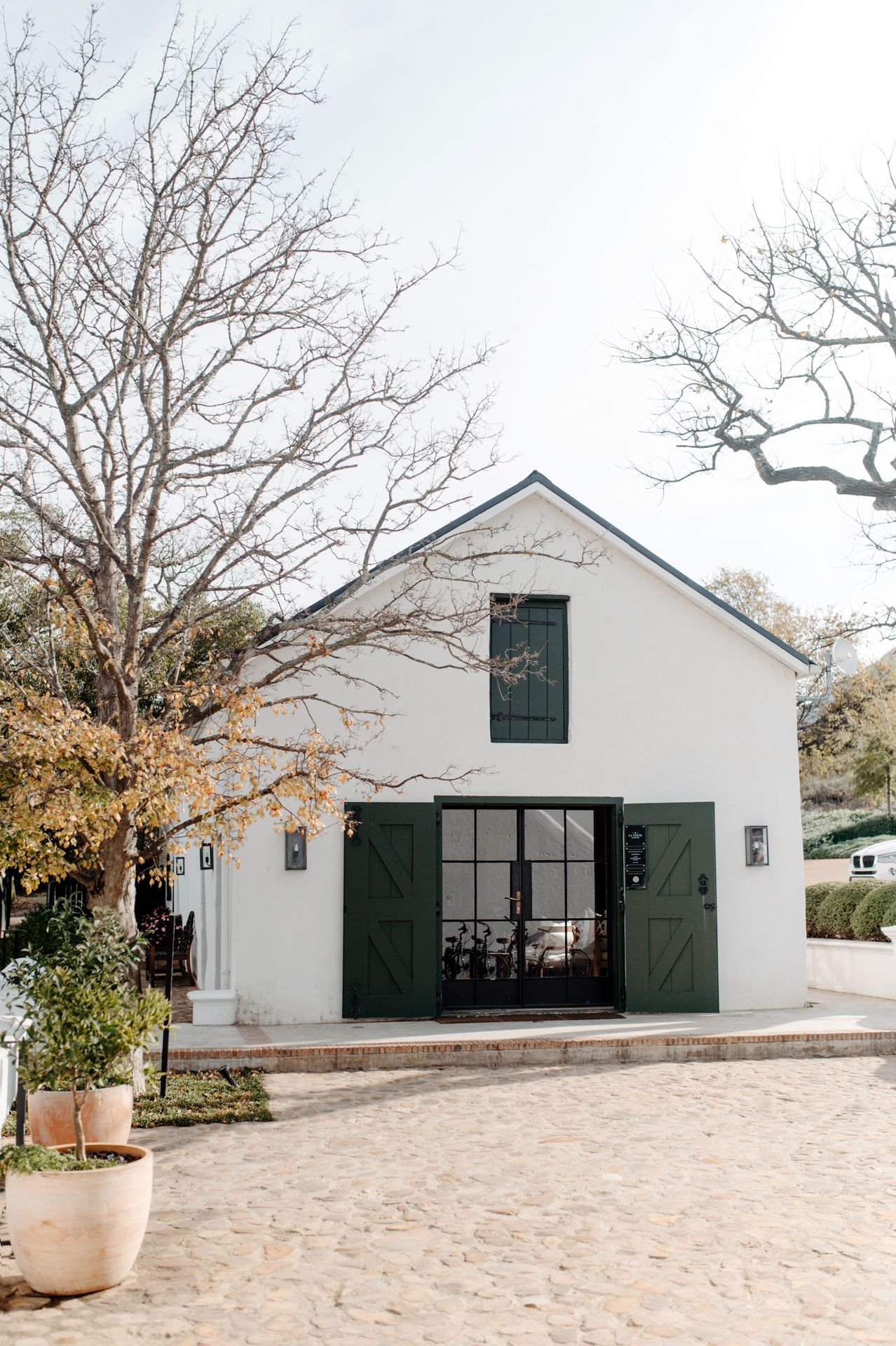 Modern white barn-style event venue with black shutters and doors set against bare winter trees and sandy ground.