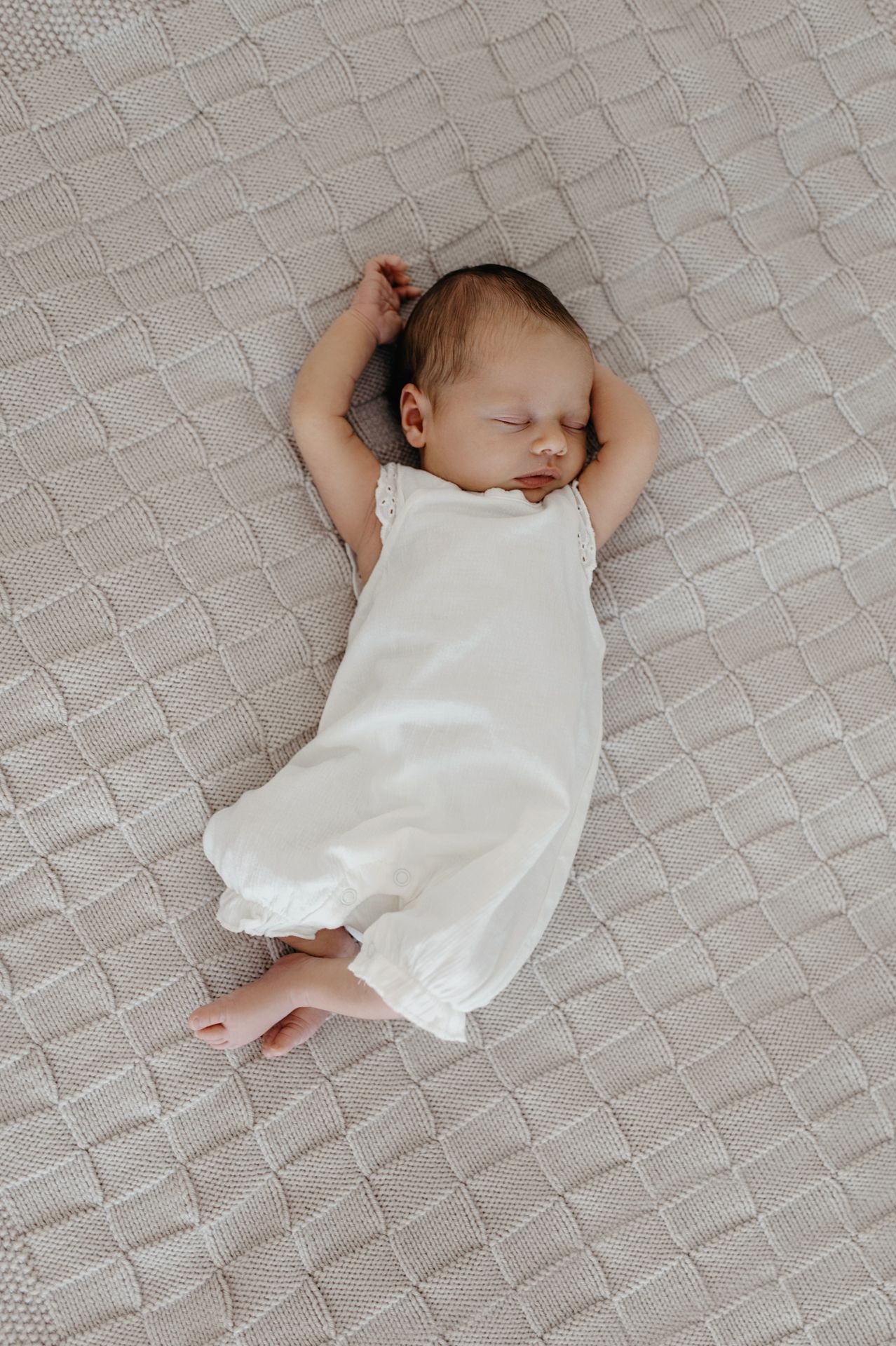 Sleeping baby in white romper lying on textured gray blanket with arms raised overhead.