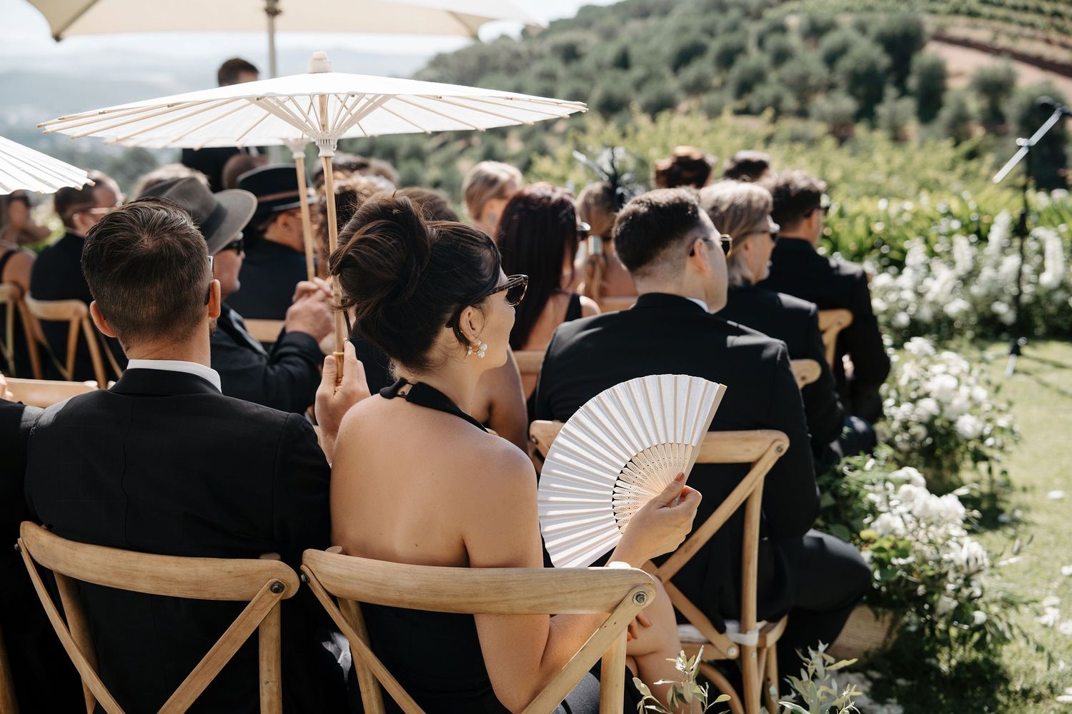 Wedding guests sit in rows under white umbrellas at an outdoor ceremony set against rolling hills and greenery.