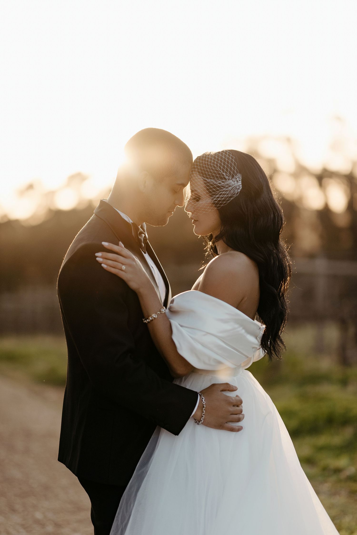 A romantic couple shares an intimate kiss at sunset in a grassy field, the bride wearing a flowing white gown.