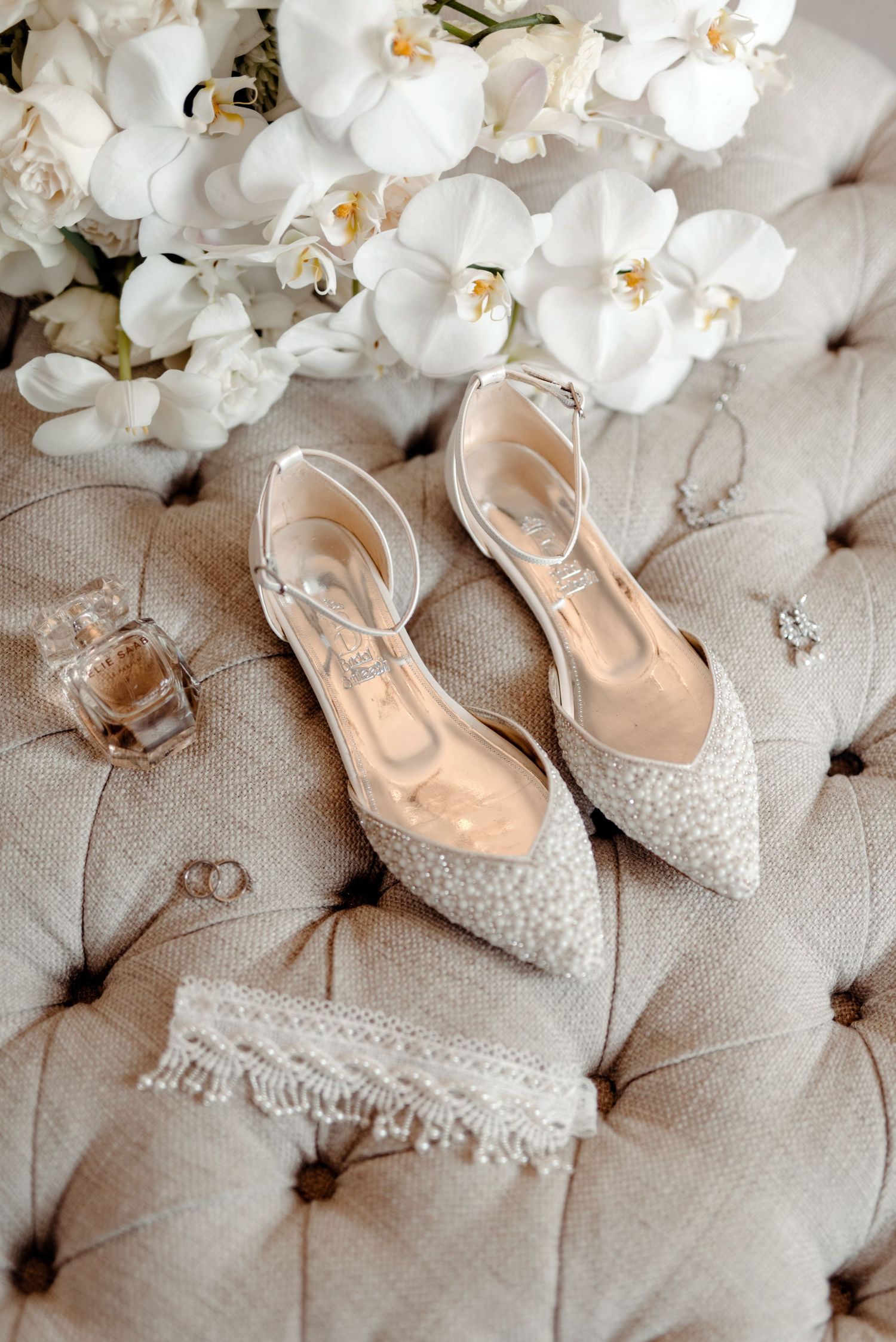White lace bridal shoes and orchids arranged on tufted upholstery with wedding rings.