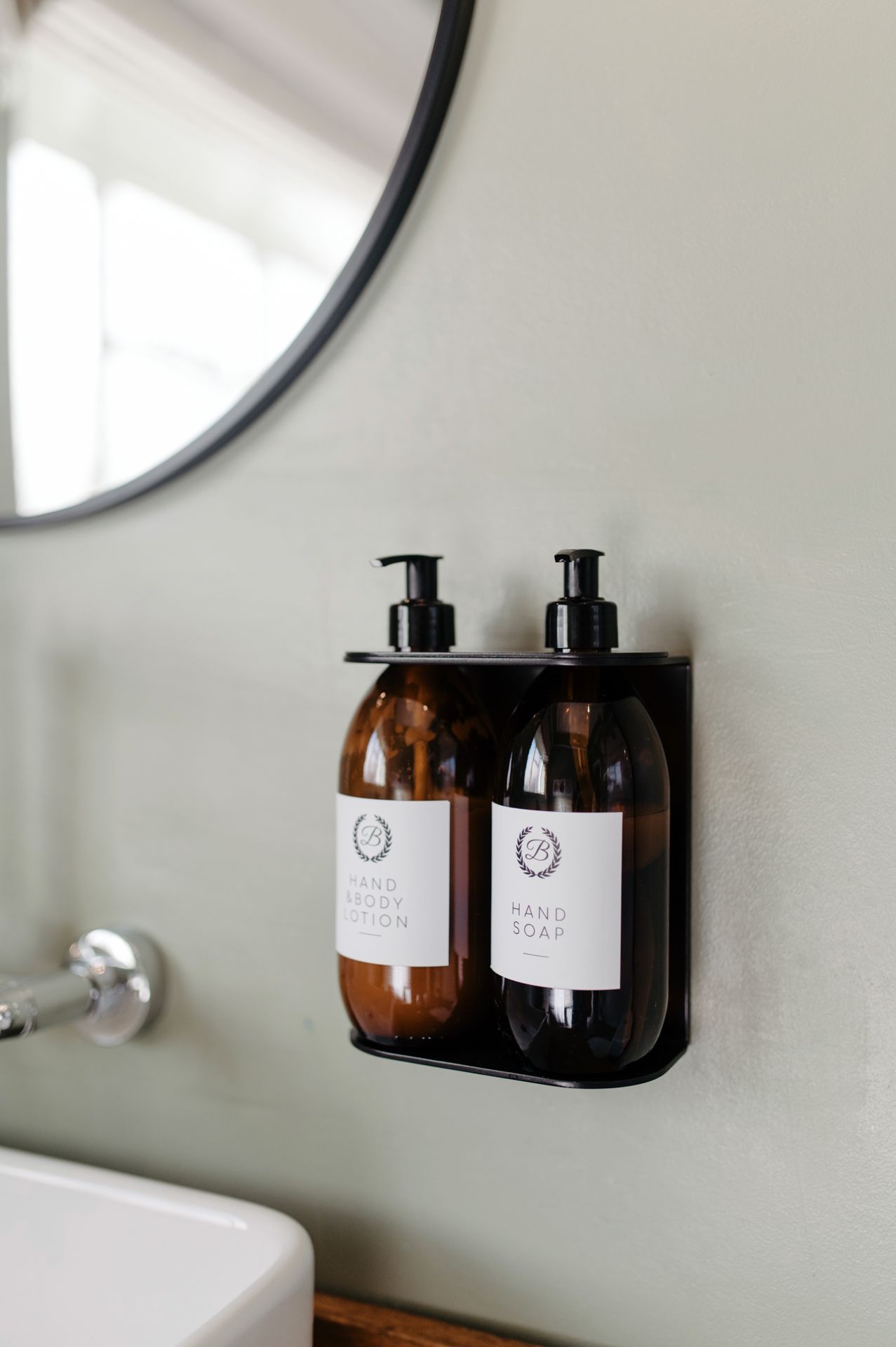 Two amber glass soap dispensers mounted on a black wall holder in a minimalist bathroom with a round mirror visible.