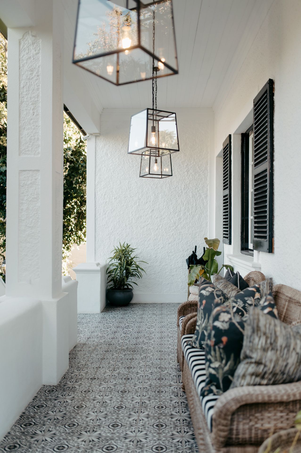 A modern covered porch features white walls, lantern pendant lights, and comfortable seating with patterned pillows.