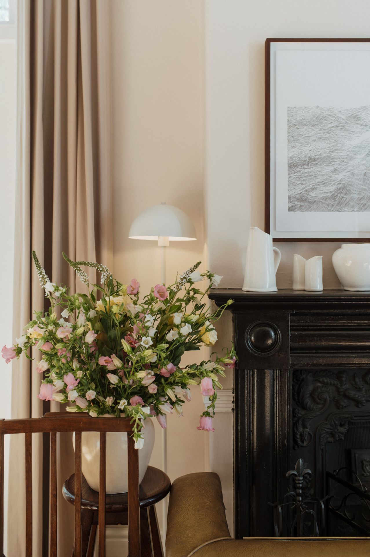 Pink and white floral arrangements in vases decorate a room with beige walls and a black fireplace mantel.