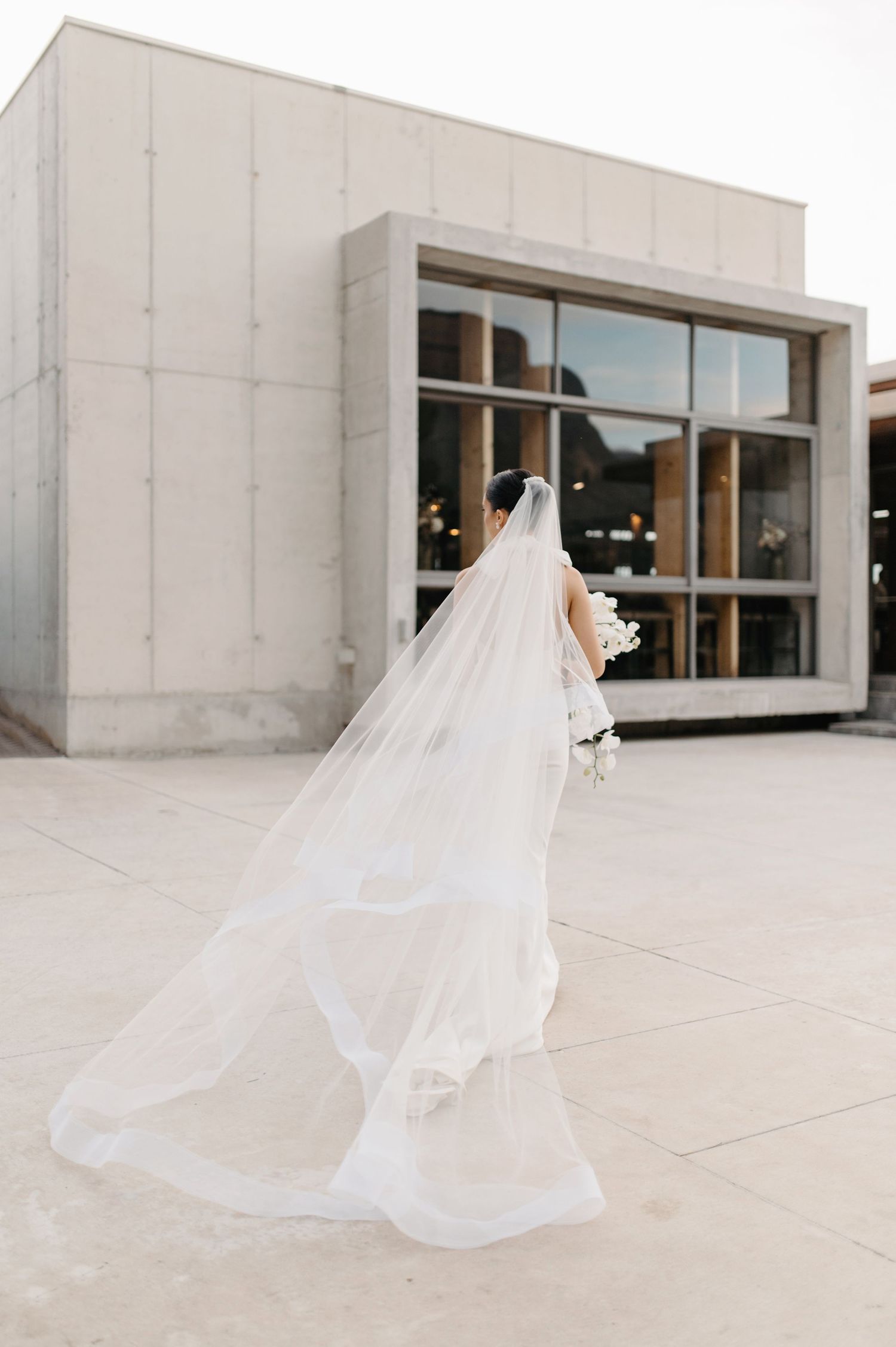 A bride in a flowing white gown with a long cathedral veil stands outside a modern building with glass windows.