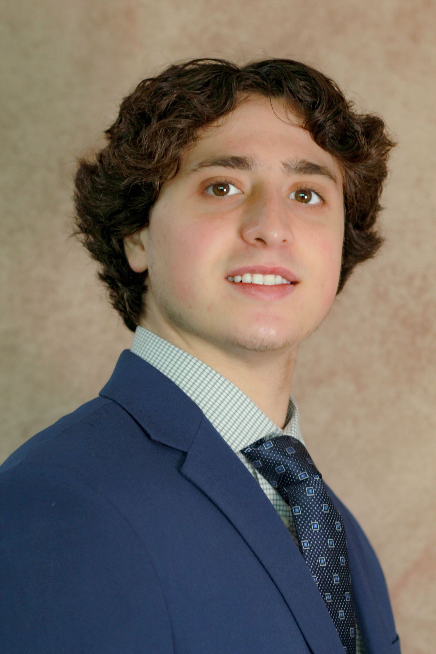 Professional headshot of a young professional in a navy suit with a patterned tie and curly hair.