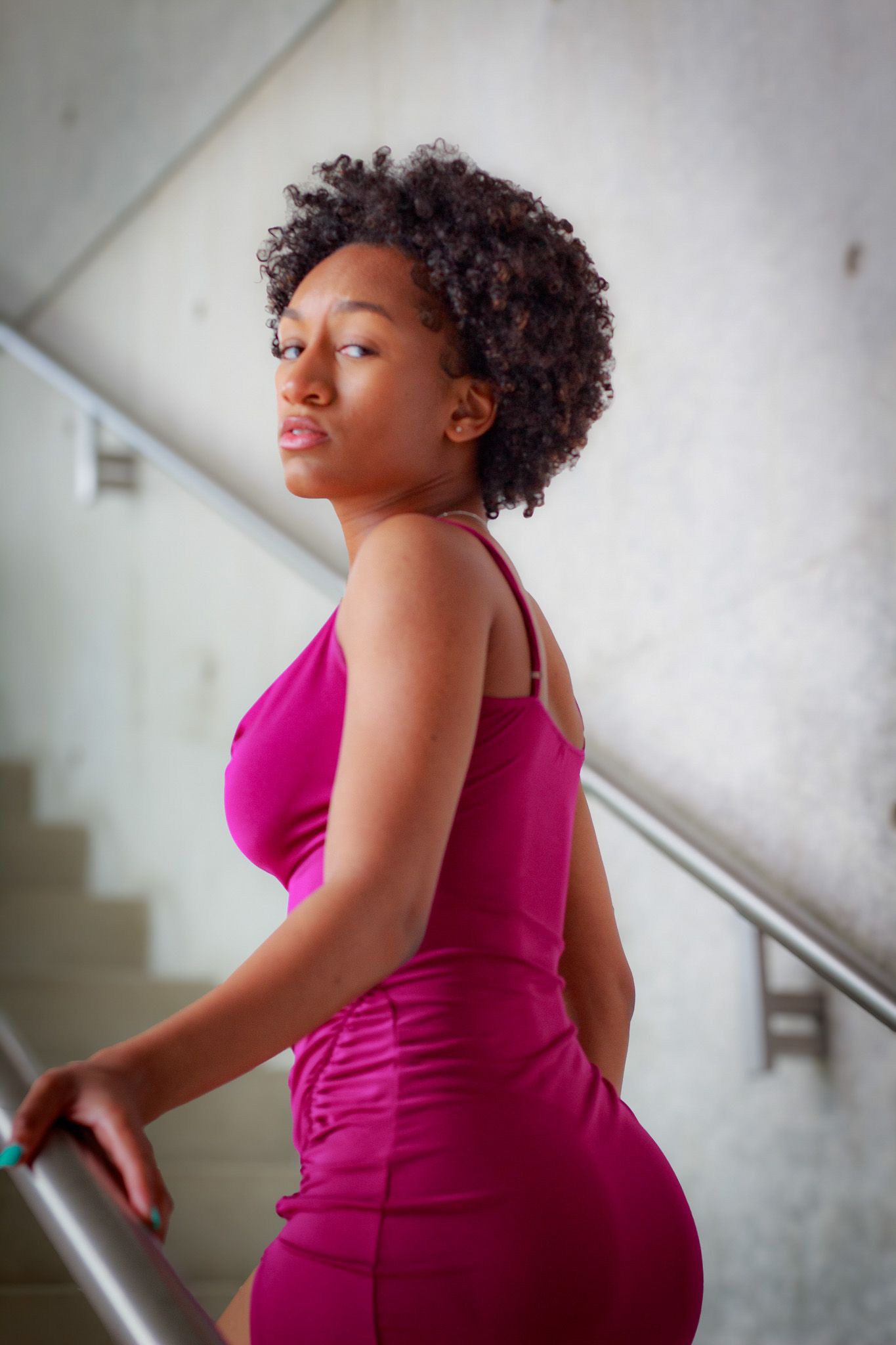 A curvy figure in a vibrant magenta dress poses confidently on stairs in an elegant indoor setting.