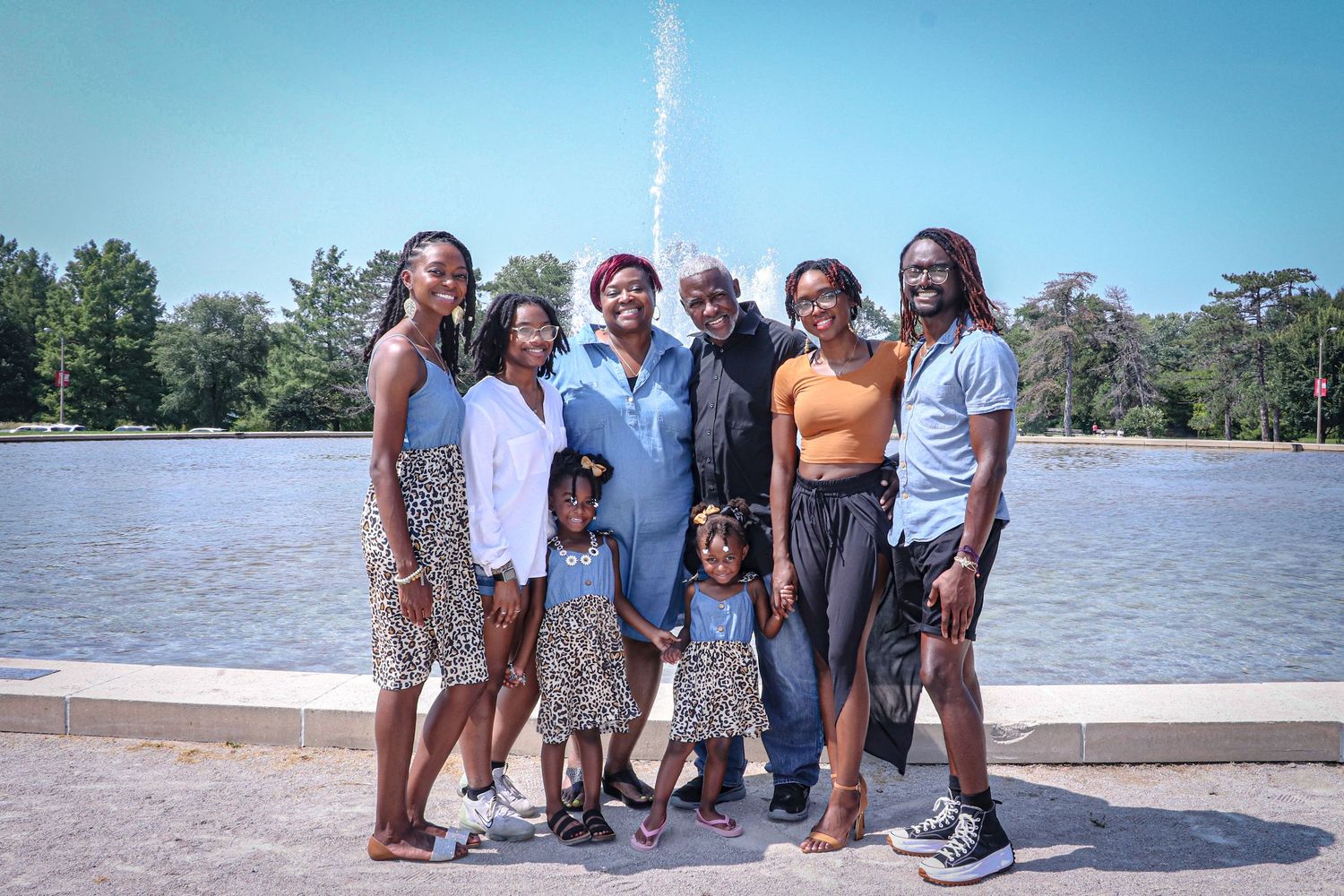 A diverse group poses together for a photo by a fountain in a sunny park setting.