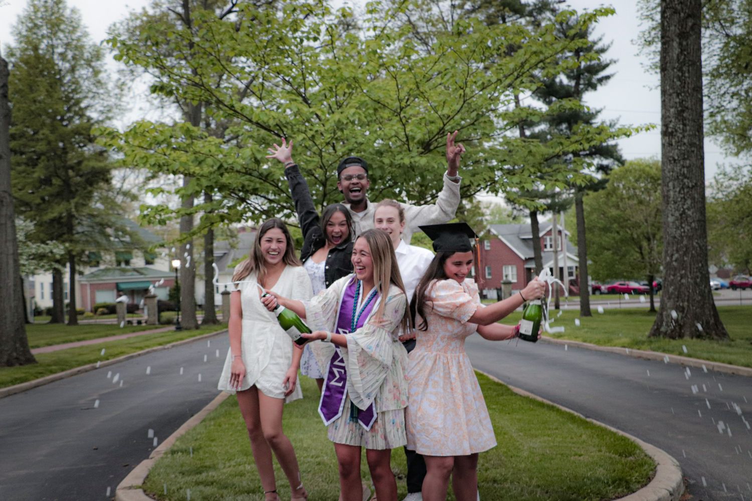 A joyful graduation celebration with friends in light dresses throwing confetti on a tree-lined street.