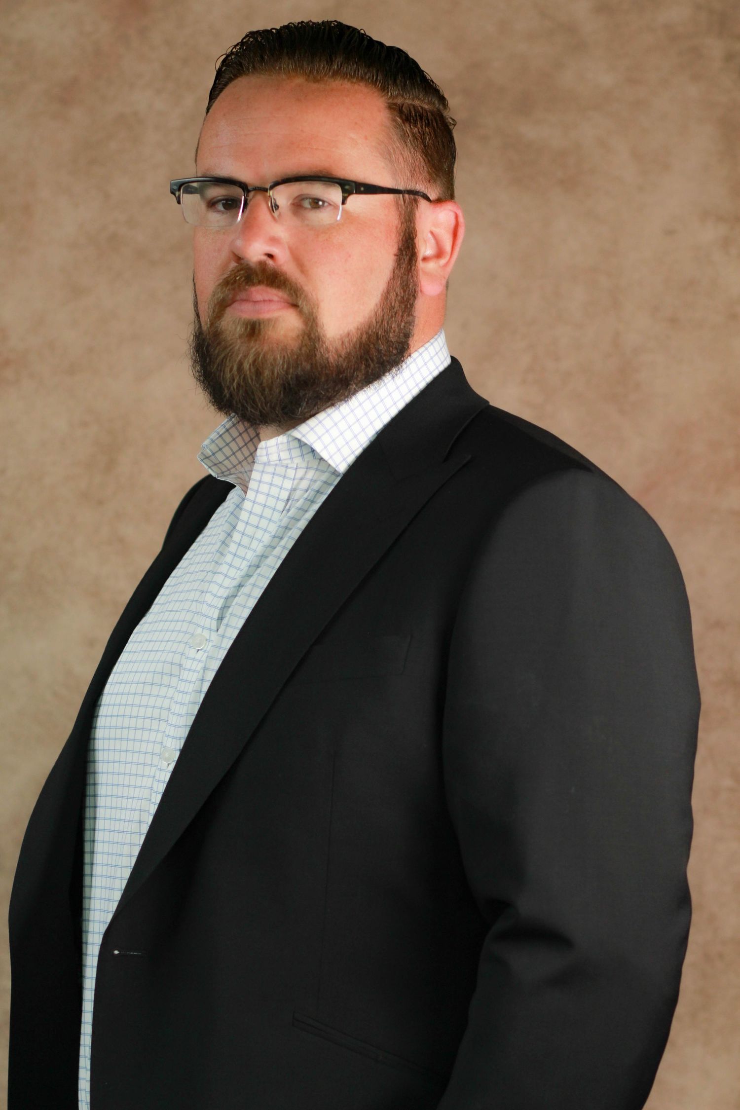 Serious looking person with beard and glasses wearing suit jacket in corporate headshot.
