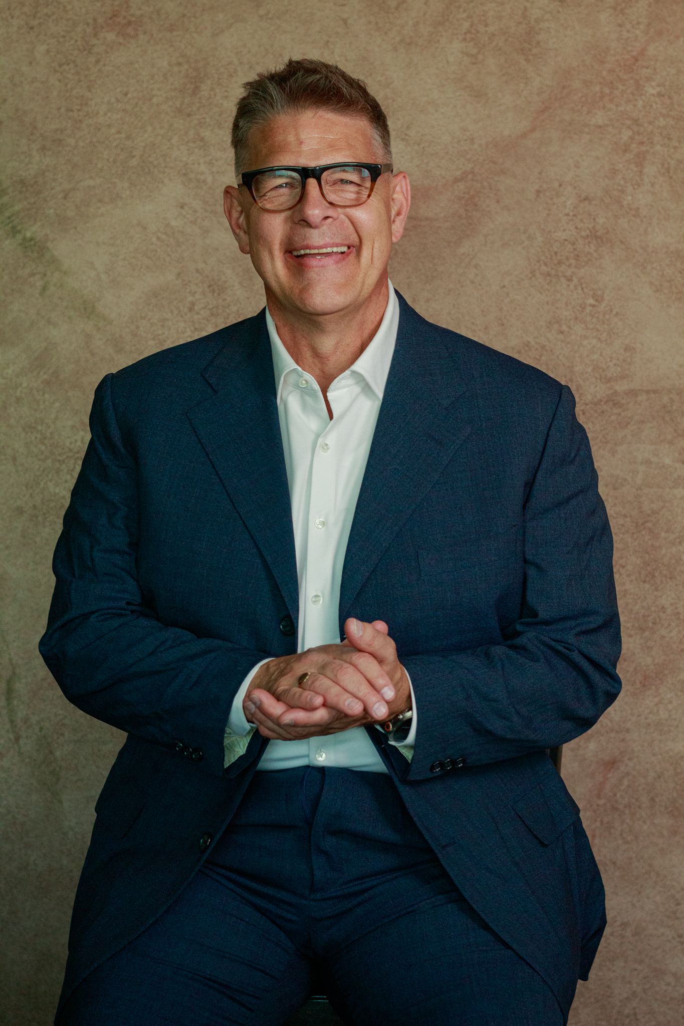 Professional portrait of a businessman in a navy suit and glasses with a warm expression.