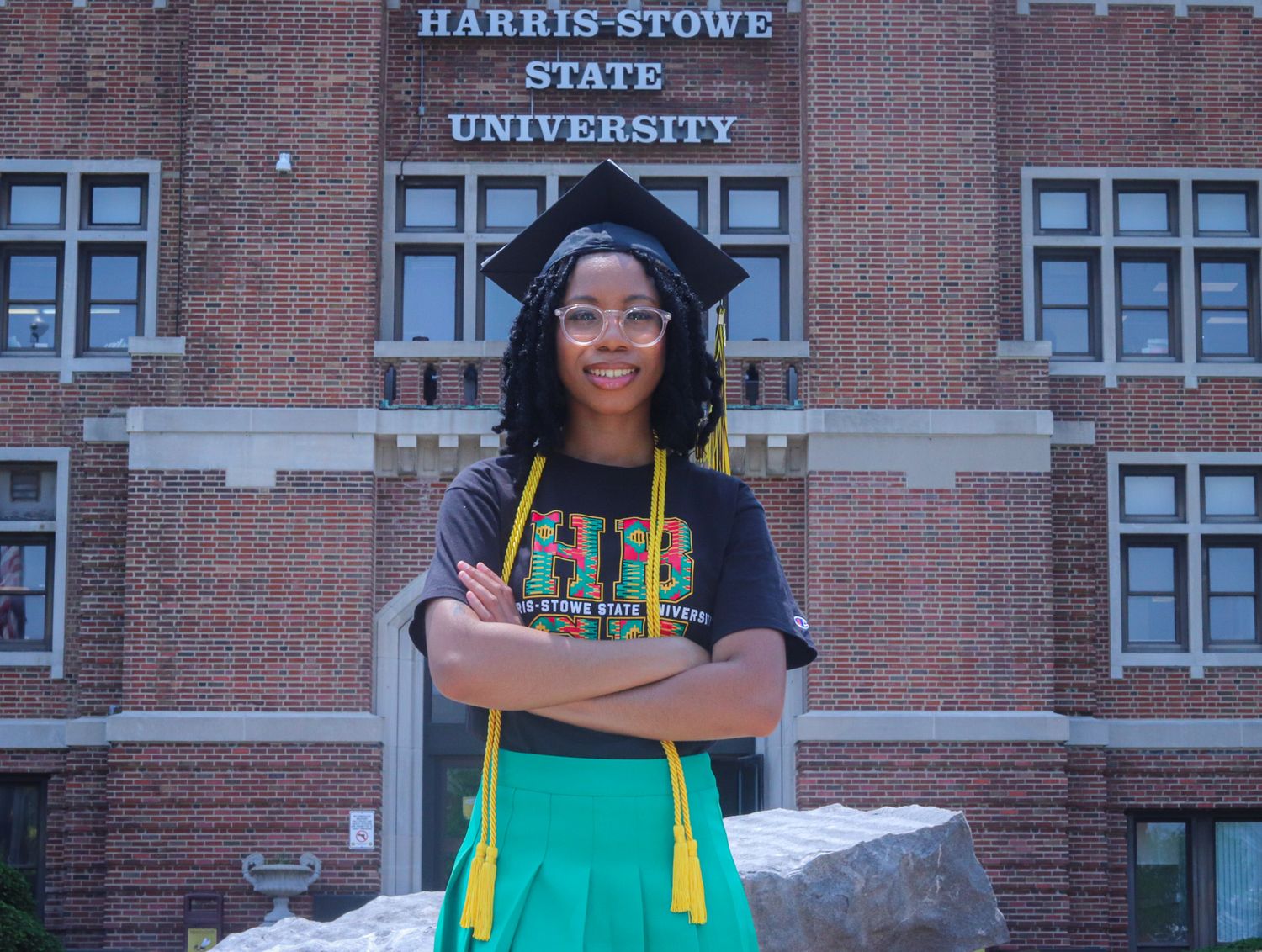 A graduate in cap and gown poses with crossed arms in front of Harris-Stowe State University building.